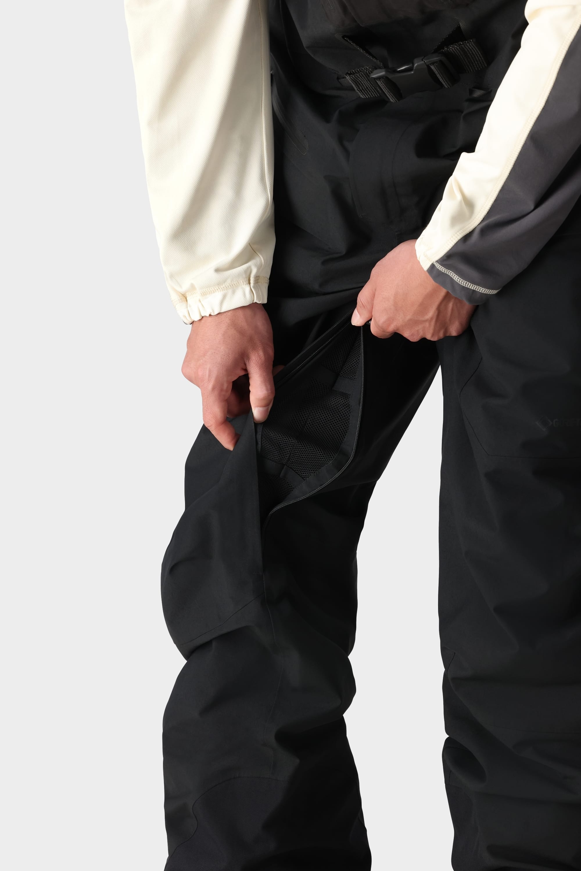 A person wearing 686 Mens GORE-TEX PRO 3L Thermagraph Bib pants is unzipping a mesh-lined vent on the side of their leg while dressed in a light-colored long sleeve shirt.