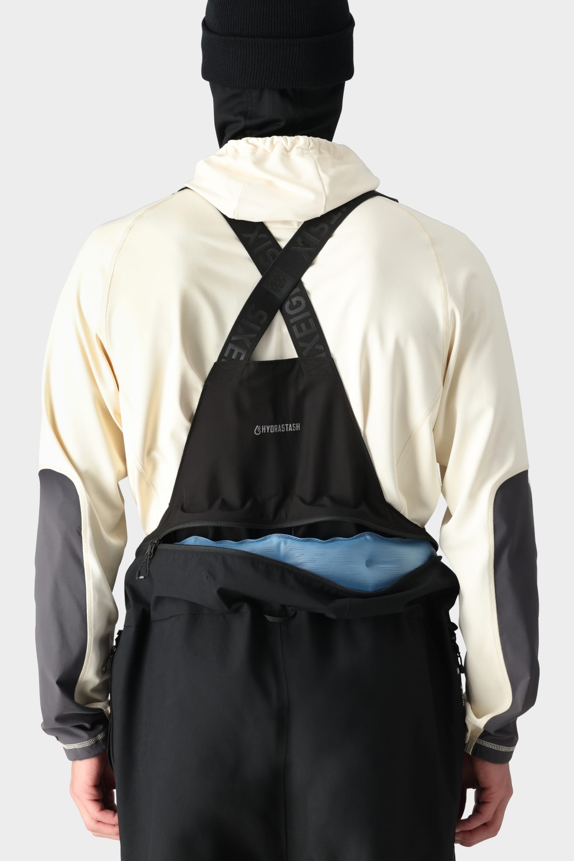 A person wearing the 686 Mens GORE-TEX PRO 3L Thermagraph Bib by 686 faces away in a cream and black jacket with gray sleeves, black cross-back suspenders, and a black beanie. A blue item is tucked into a waist pouch.