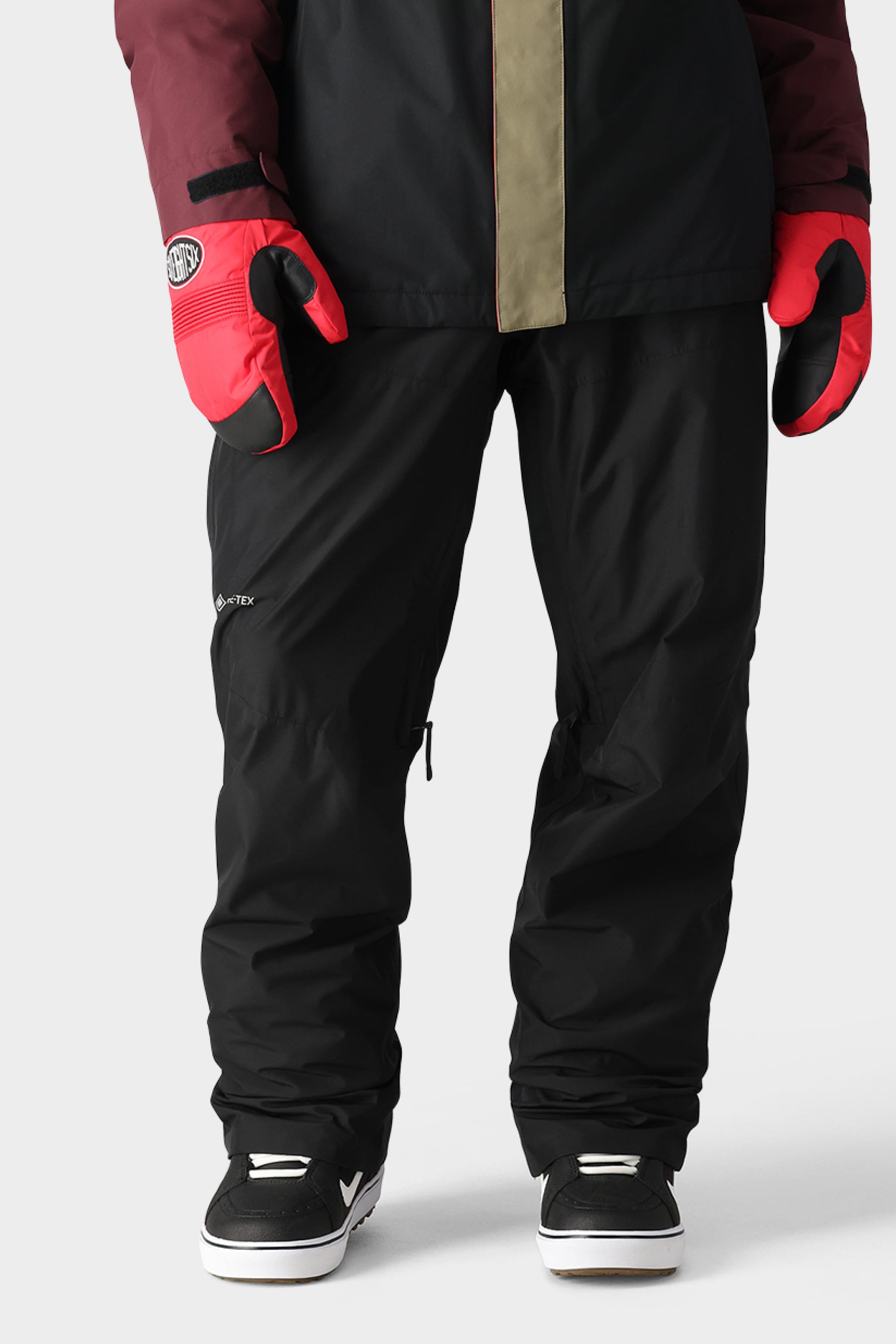 A person wearing 686 Mens GORE-TEX Core Shell Pant by 686, black and white sneakers, a dark jacket with an olive vertical stripe, and red mittens with black accents stands against a plain light background.