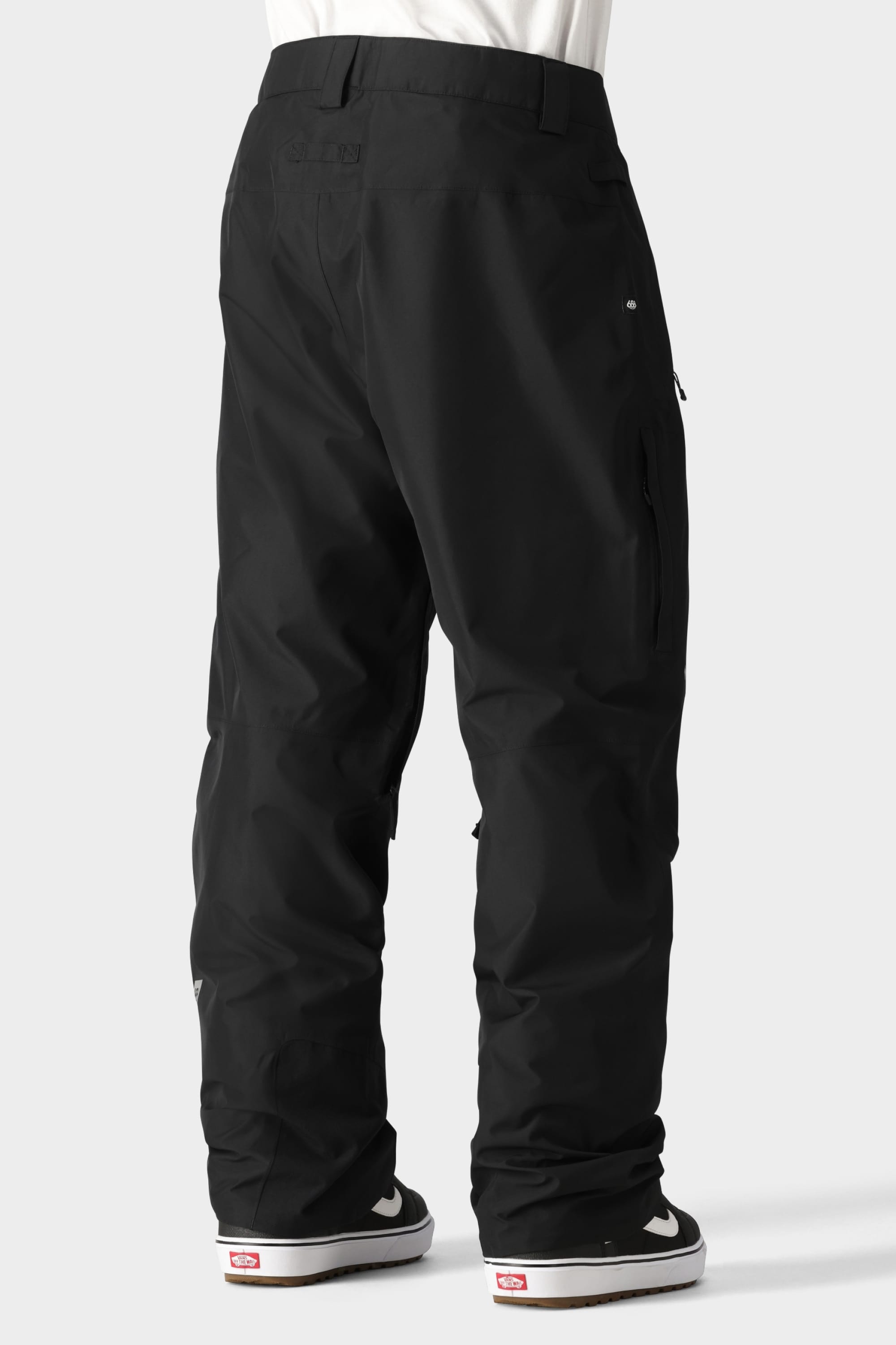 A person, shown from the waist down against a light gray background, wears 686 Mens GORE-TEX Core Shell Pants in black and black-and-white sneakers with red logos.