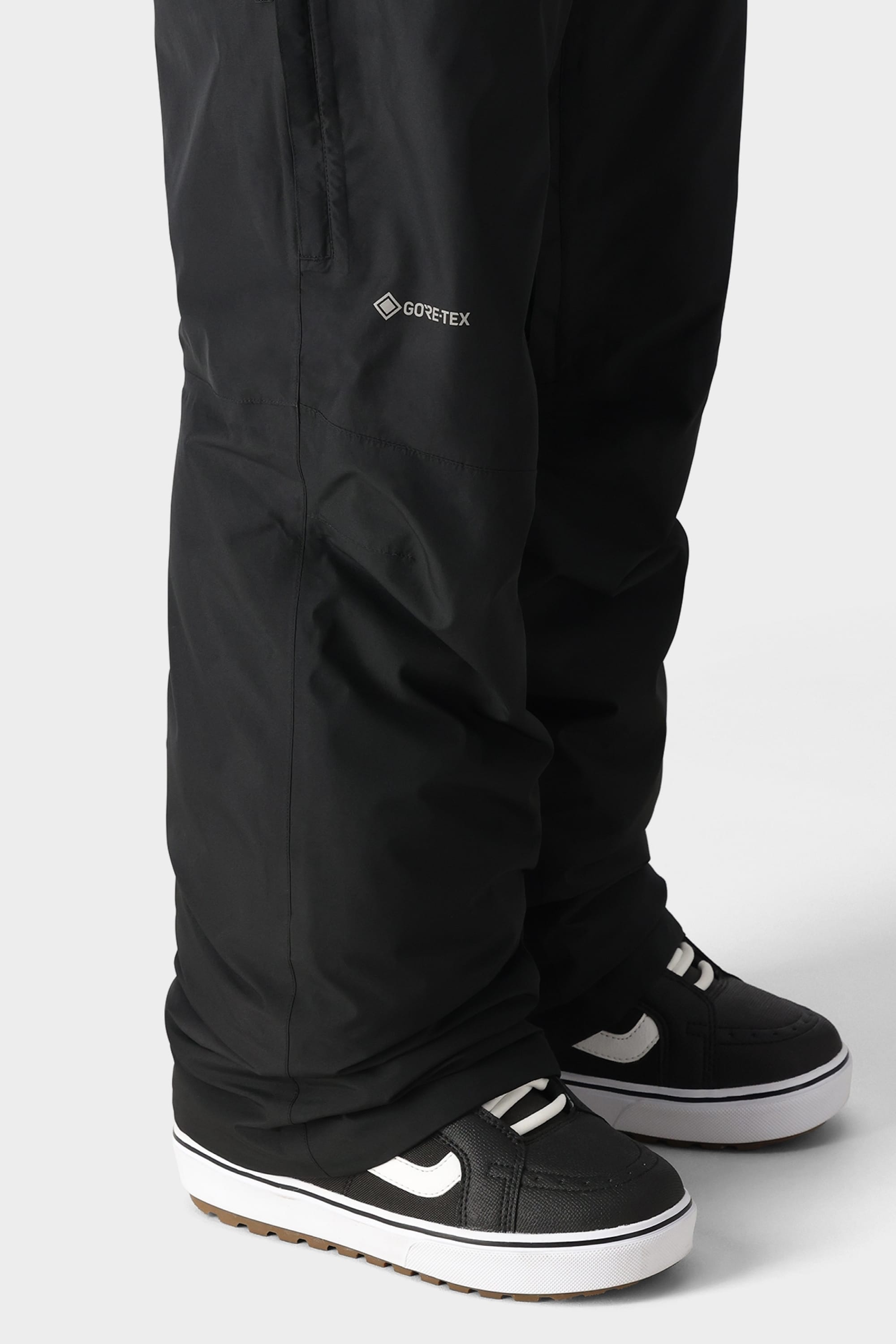 A person in 686 Mens GORE-TEX Core Shell Pants and black-and-white sneakers stands on a light gray surface, with only their lower legs and feet visible.