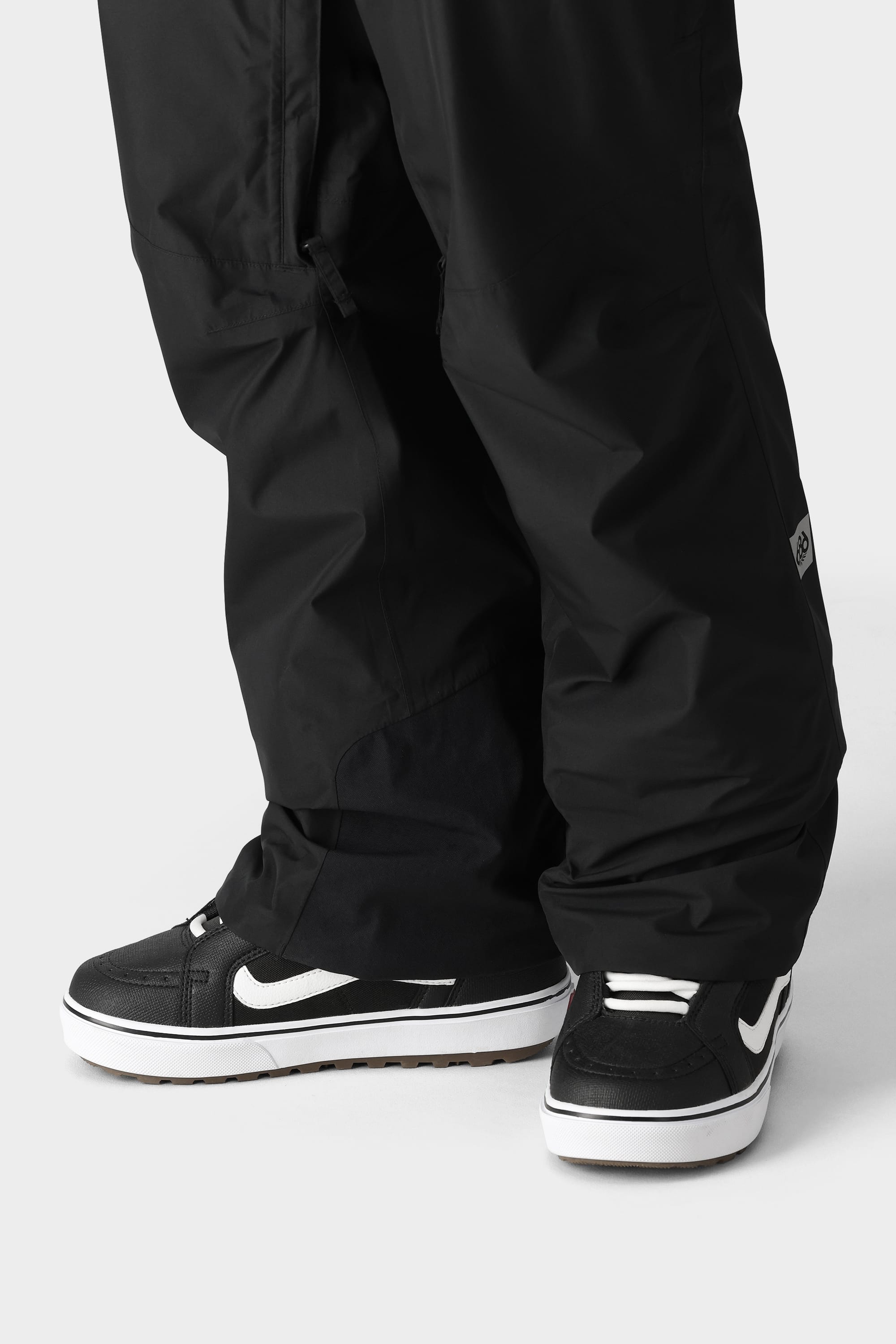 A person wears 686 Mens GORE-TEX Core Shell Pants in black—durable, breathable, and loose-fit—paired with black-and-white Nike sneakers, standing on a light gray surface with pants partially covering the shoes.