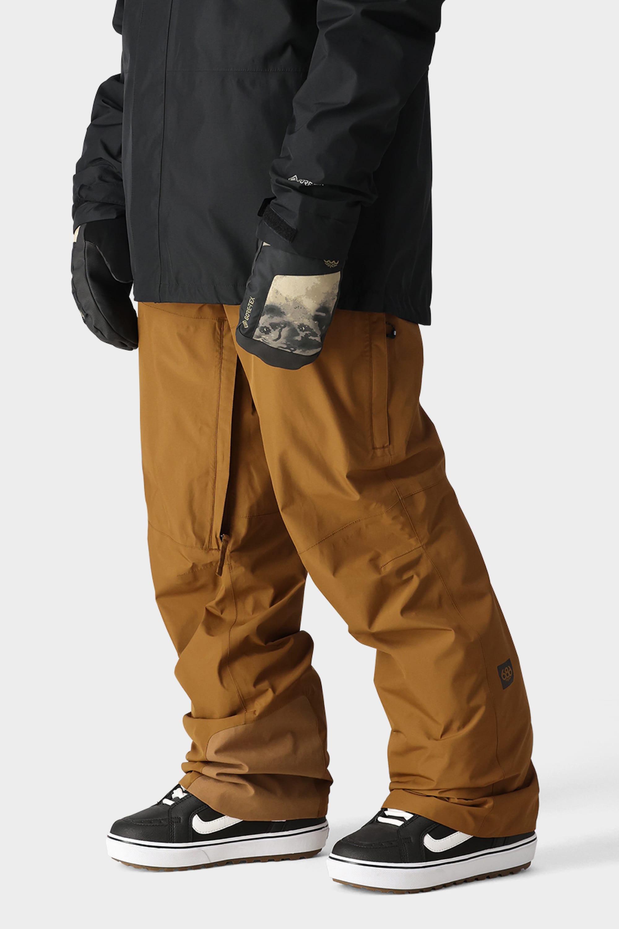 A person wears durable brown 686 Men’s GORE-TEX Core Shell Pant, a black jacket, black gloves with a graphic print, and black Nike sneakers with white soles, standing against a plain light background.
