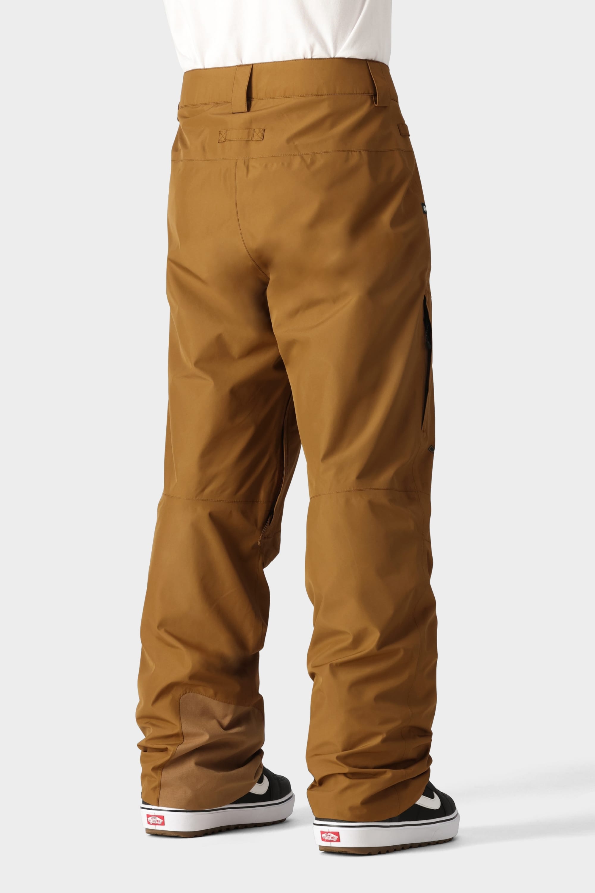 Model wearing 686 Mens GORE-TEX Core Shell Pant in brown—breathable and durable—with black and white sneakers, shown from the back against a plain light gray background.