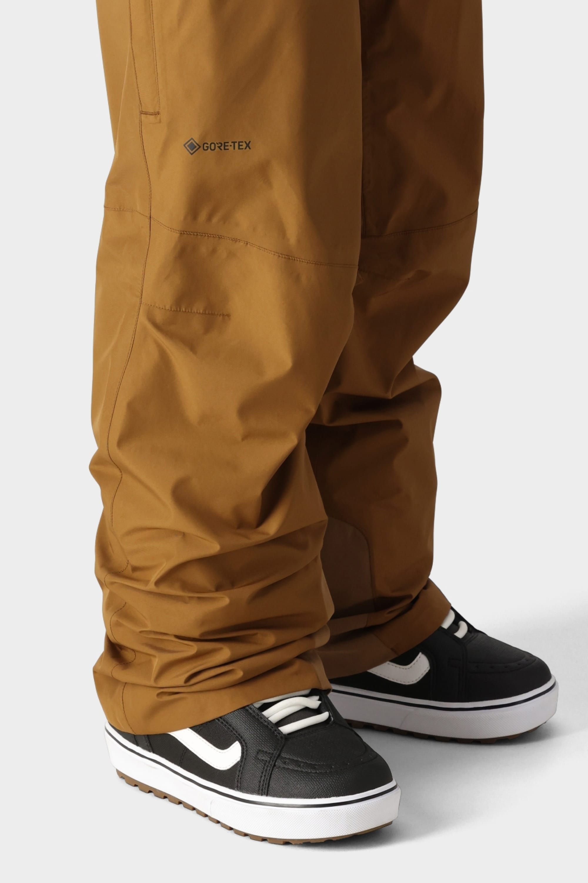Someone wearing 686 Mens GORE-TEX Core Shell Pants in brown with black and white Vans sneakers, shown from the knee down against a plain light background.