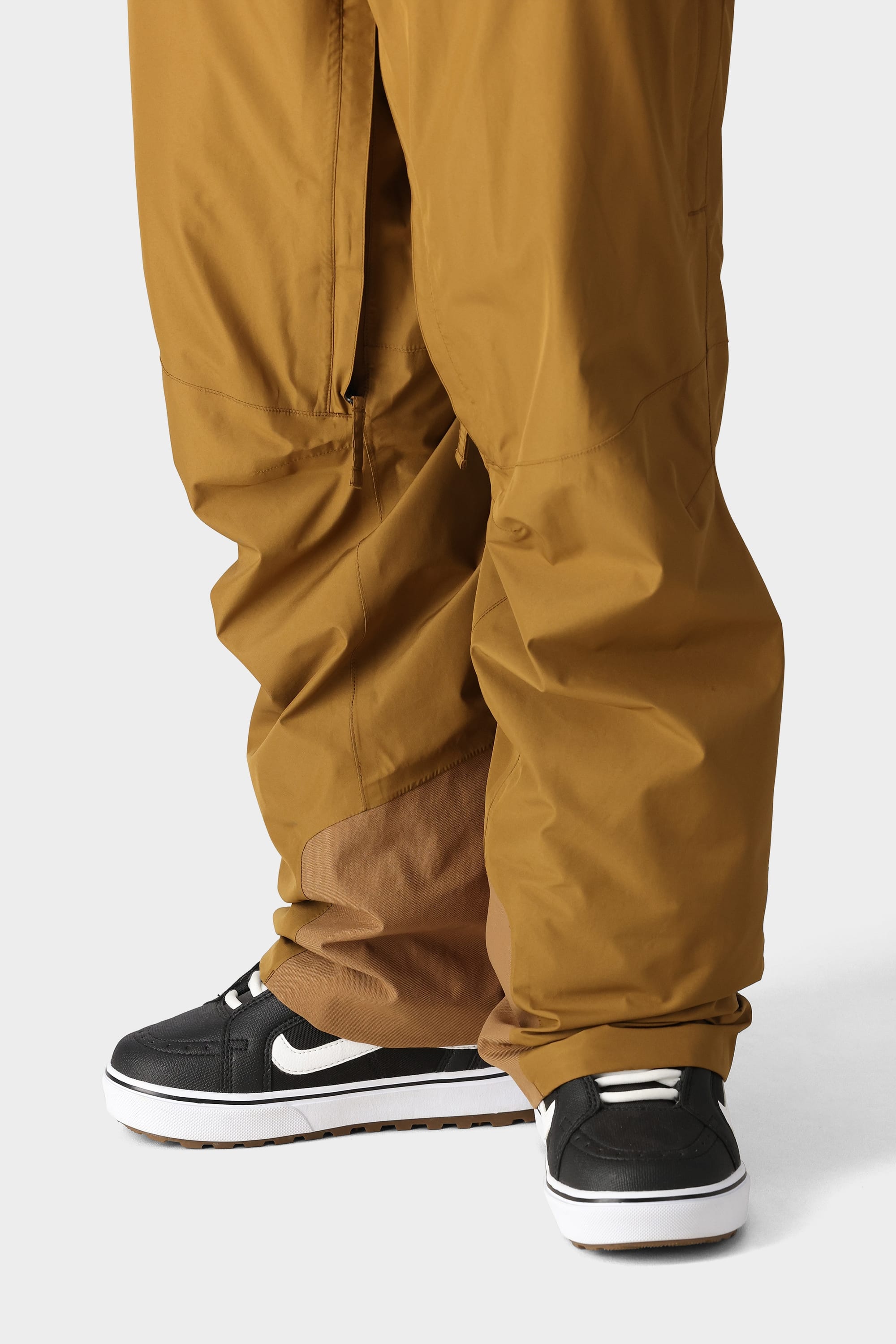 A person wearing 686 Mens GORE-TEX Core Shell Pant by 686 in brown, paired with black-and-white Nike sneakers, stands on a light gray surface with the pants slightly covering the shoes.