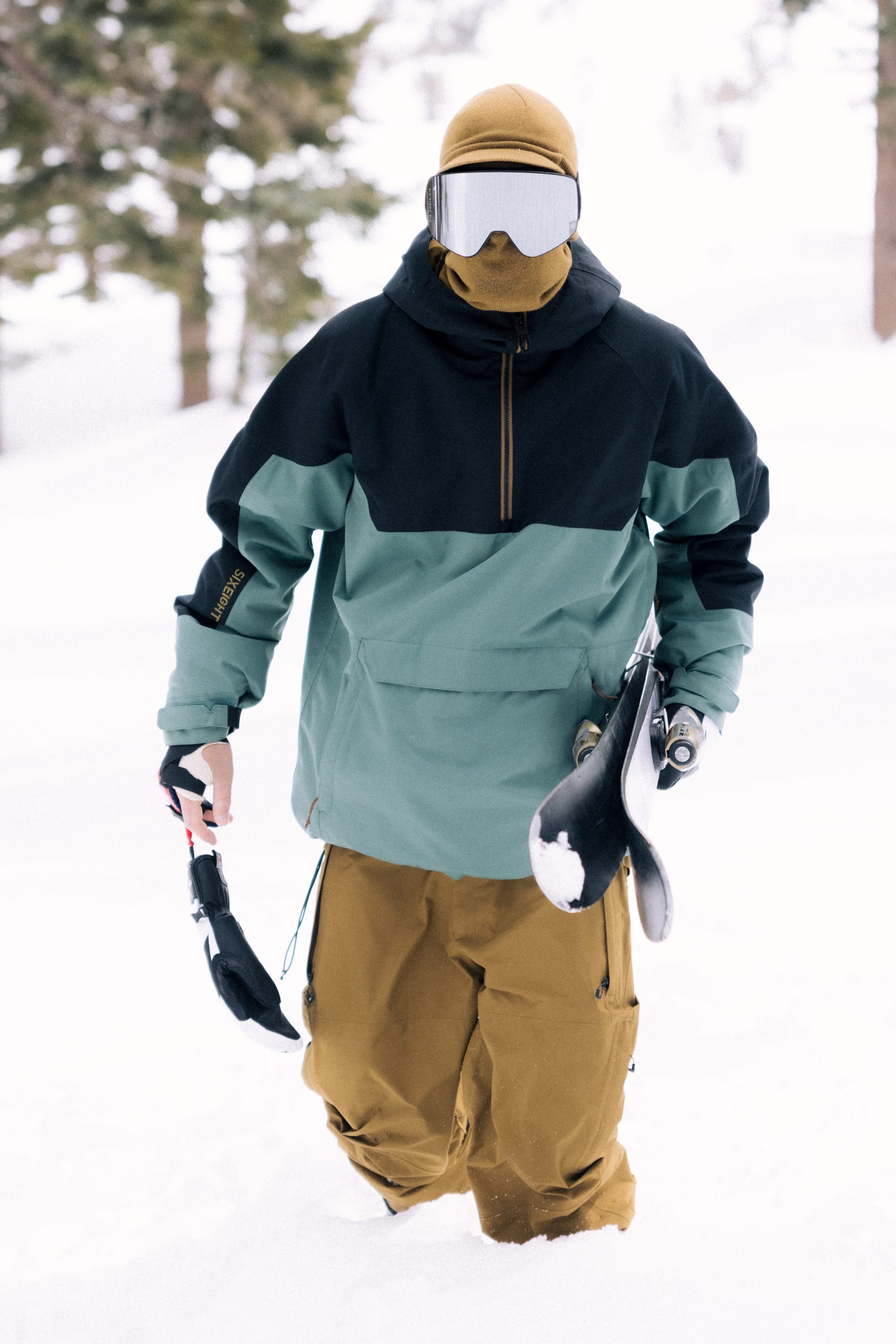 Wearing the 686 Mens GORE-TEX Core Shell Pant, a person in snow gear with a tan hood, goggles, and a teal and black jacket stands in deep snow holding a snowboard, with snowy trees in the background.