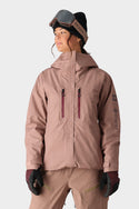 A woman stands against a plain light background wearing the 686 Womens GORE-TEX Skyline Shell Jacket in light brown, with matching pants, a black beanie, ski goggles on her forehead, and black gloves.