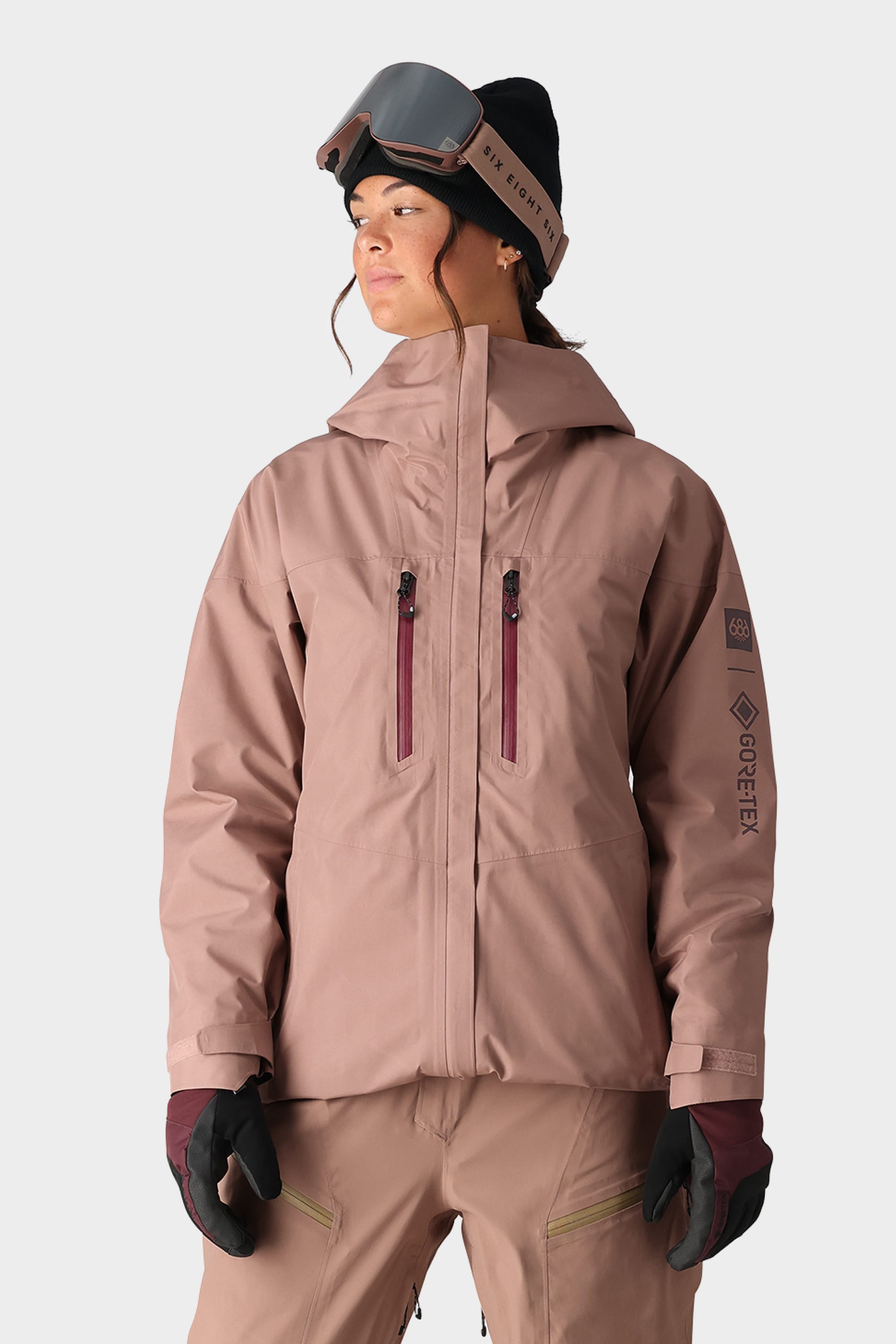 A woman stands against a plain light background wearing the 686 Womens GORE-TEX Skyline Shell Jacket in light brown, with matching pants, a black beanie, ski goggles on her forehead, and black gloves.