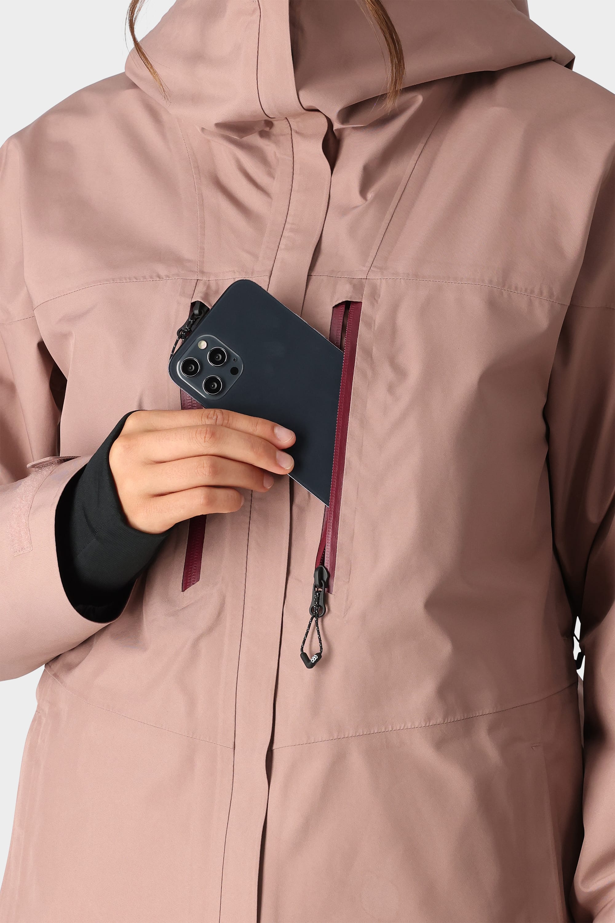 A woman in the 686 Womens GORE-TEX Skyline Shell Jacket holds a smartphone partially inside a zipped chest pocket; only her lower face and hand are visible.