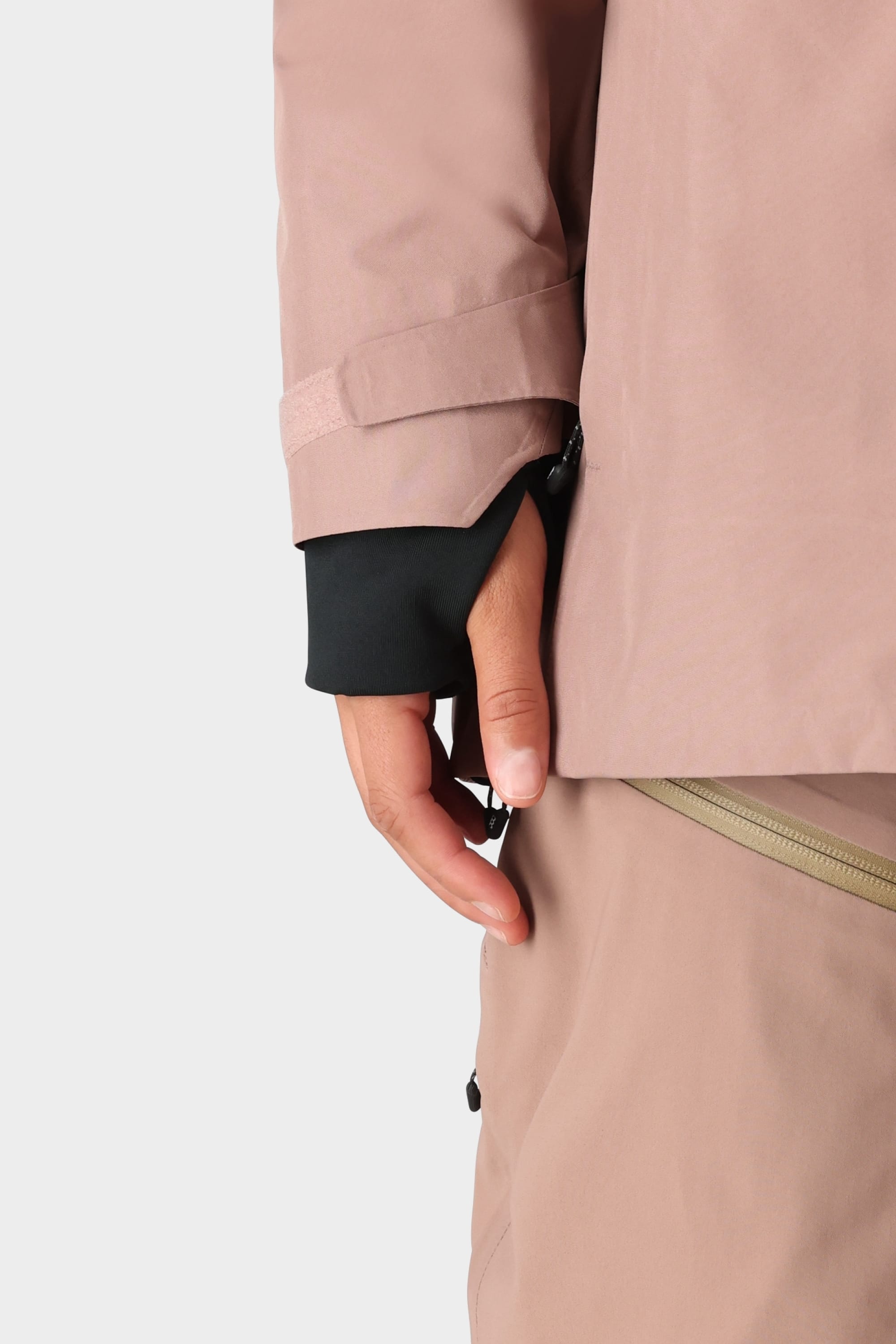 A person wears the 686 Womens GORE-TEX Skyline Shell Jacket in light pink with matching pants, a black inner sleeve visible at the wrist, hand partially tucked into the sleeve, and a zipper detail on the pants.