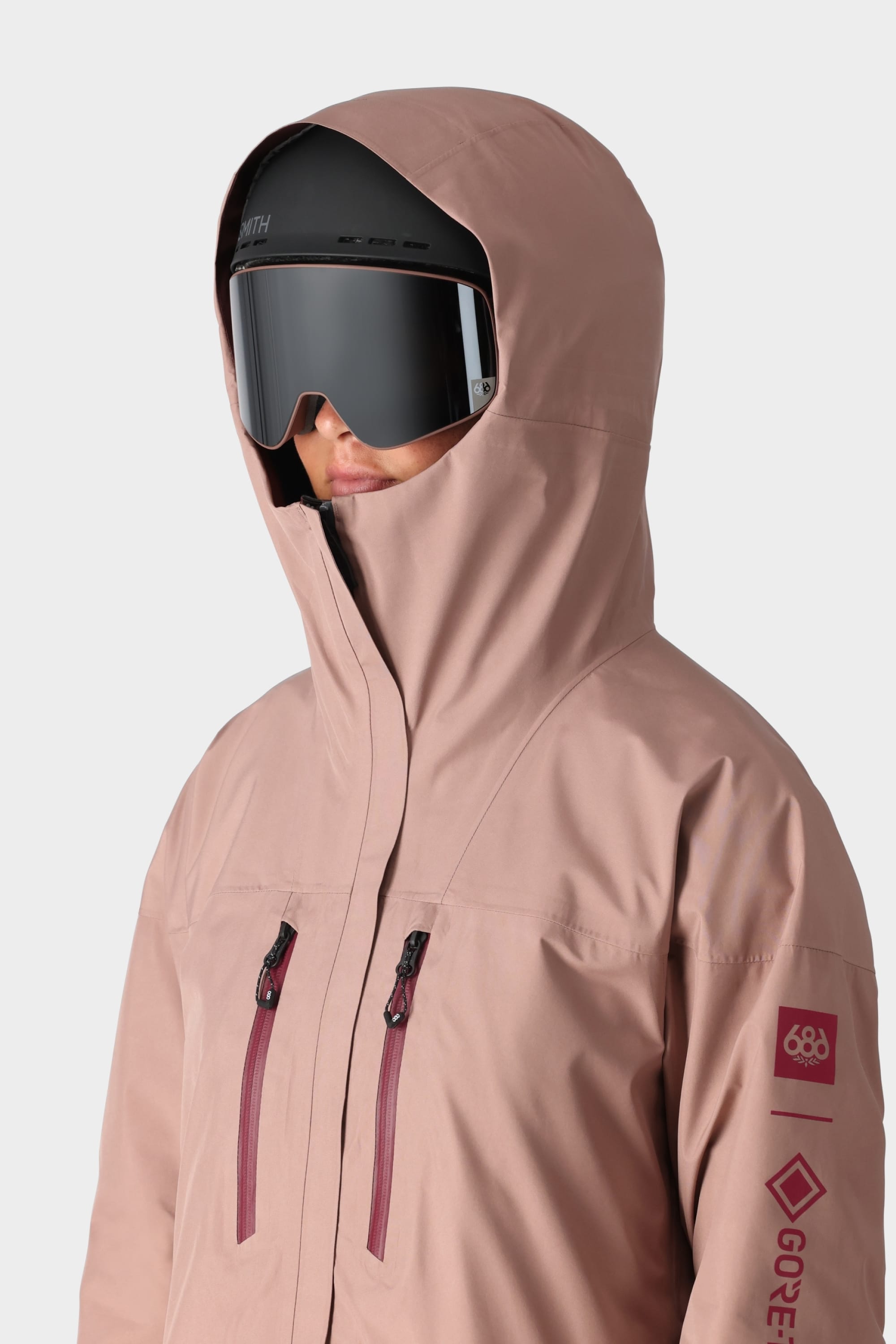 A person wears the 686 Womens GORE-TEX Skyline Shell Jacket in pink with maroon zippers, plus black ski goggles and a helmet. The 686 and GORE-TEX logos appear on the sleeve. The background is plain white.