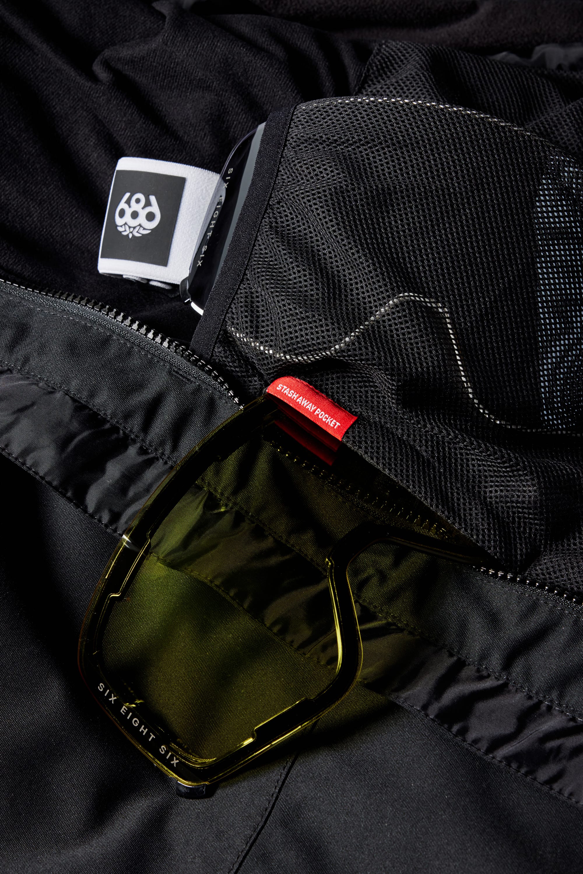 A close-up of black mesh and fabric on the 686 Womens GORE-TEX Skyline Shell Jacket, featuring a red SIXEIGHTSEVENPROJECT tag, yellow-tinted sunglasses in a zippered pocket, and a white 686 logo atop this stormproof shell with body-mapped lining.