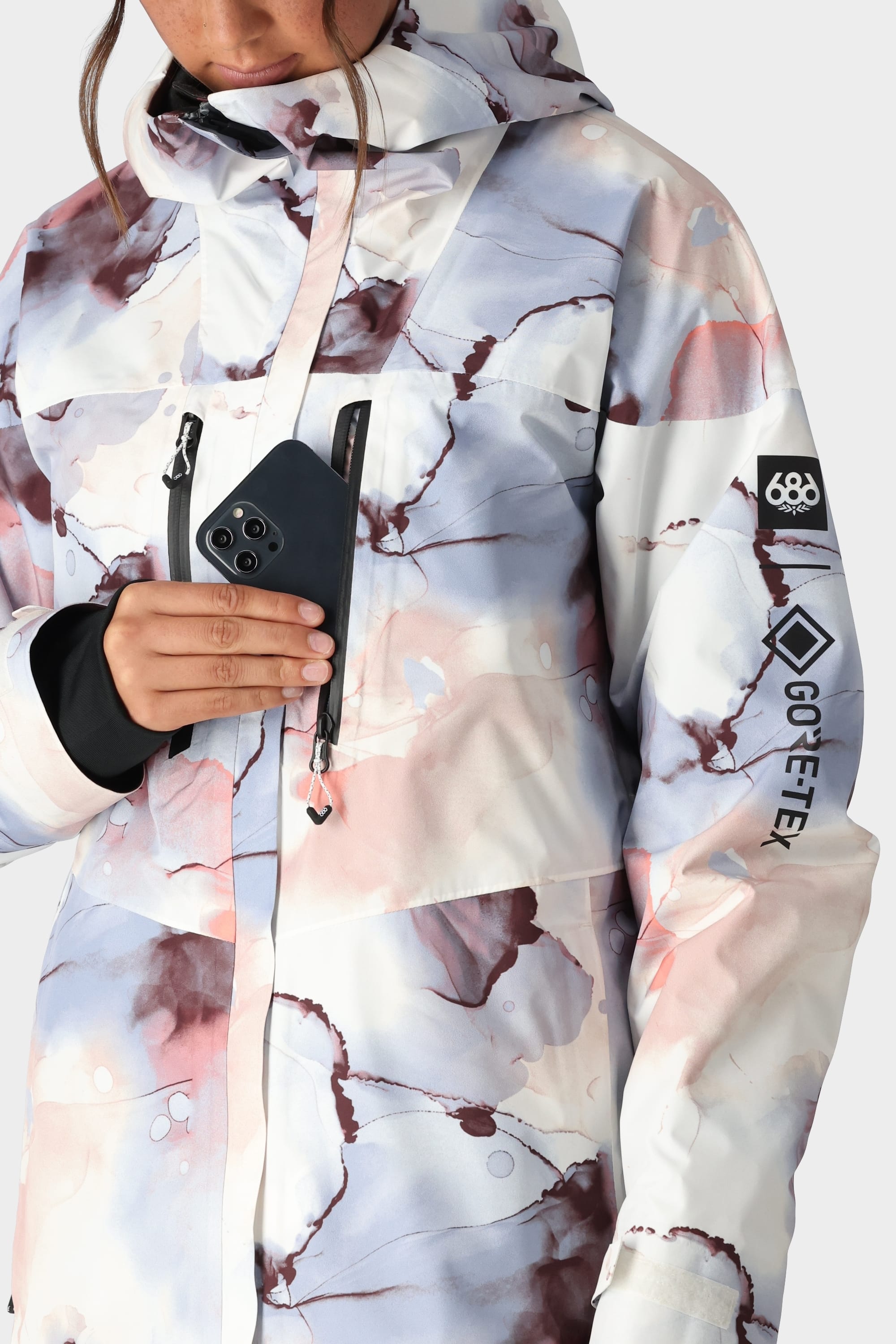A person in the 686 Womens GORE-TEX Skyline Shell Jacket, featuring a pastel marble print, hood, stormproof shell, body-mapped lining, black inner cuffs, and GORE-TEX logos, slips a black smartphone into the chest pocket.