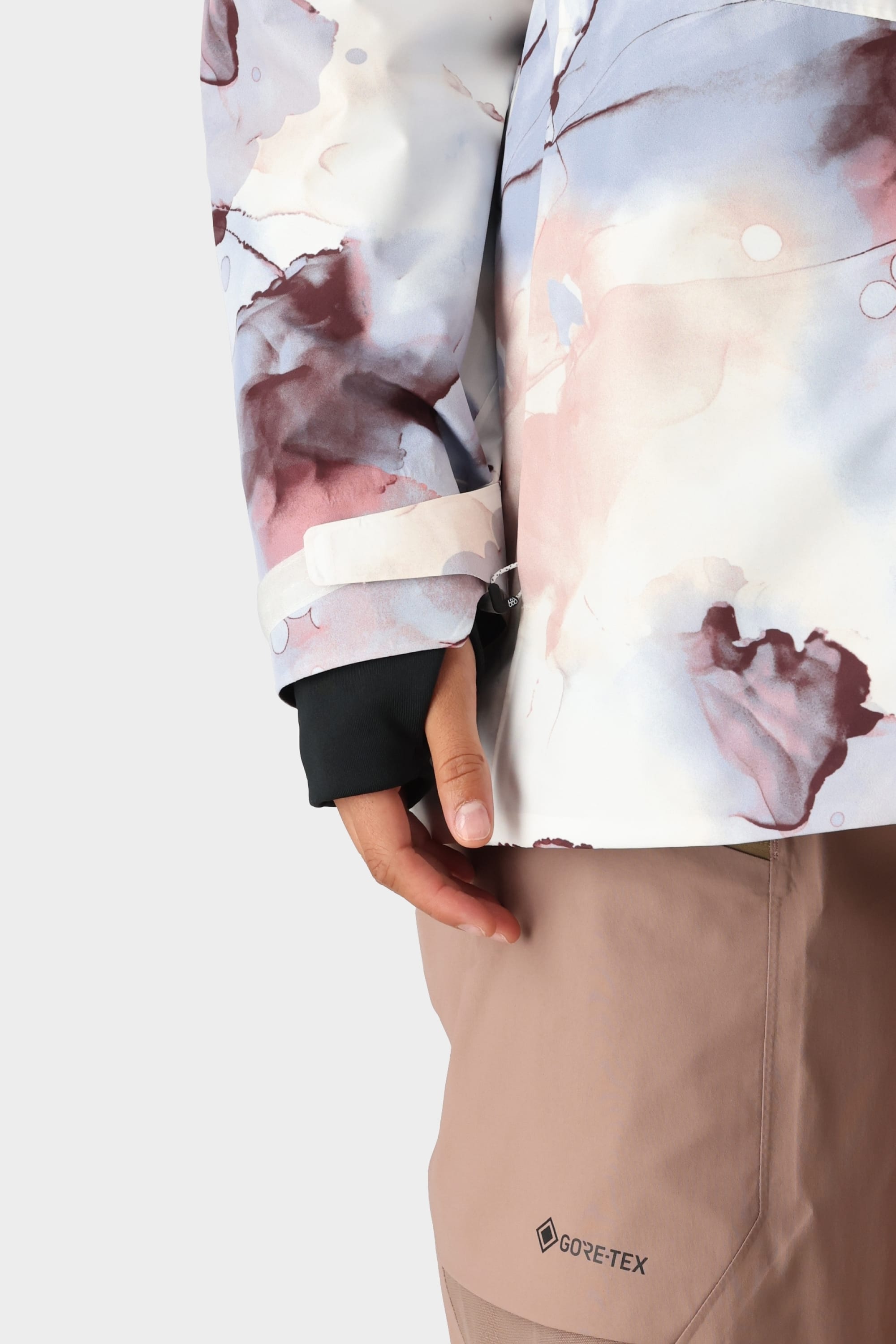 A person in the 686 Womens GORE-TEX Skyline Shell Jacket with an abstract pastel design and light brown GORE-TEX pants, showing only a lower arm, hand, and parts of the jacket and pants against a plain background.
