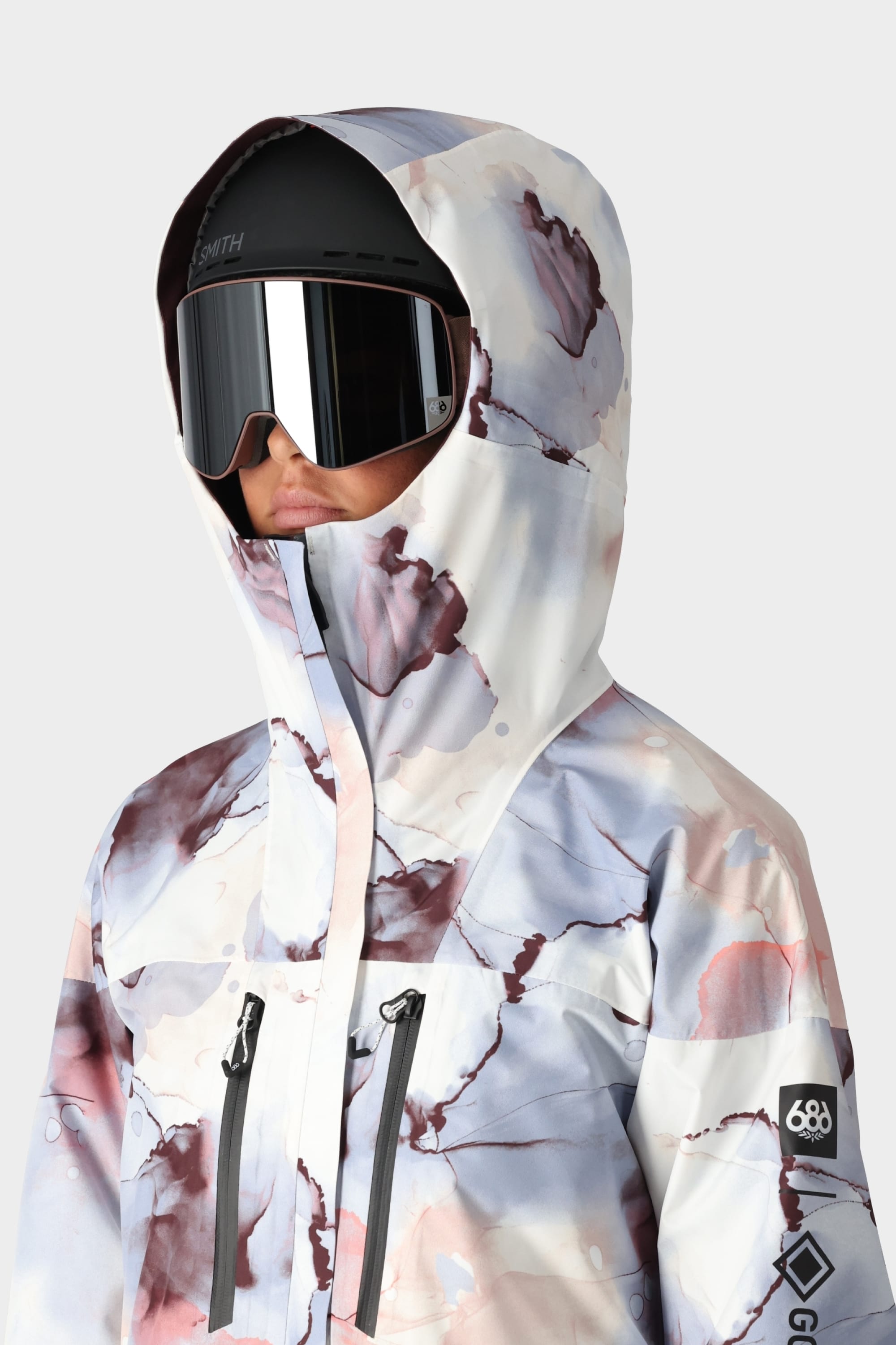 A person wears the 686 Womens GORE-TEX Skyline Shell Jacket with abstract pastel patterns, black zipper pockets, a 686 logo on the left sleeve, large ski goggles, and body-mapped lining. The background is plain light gray.