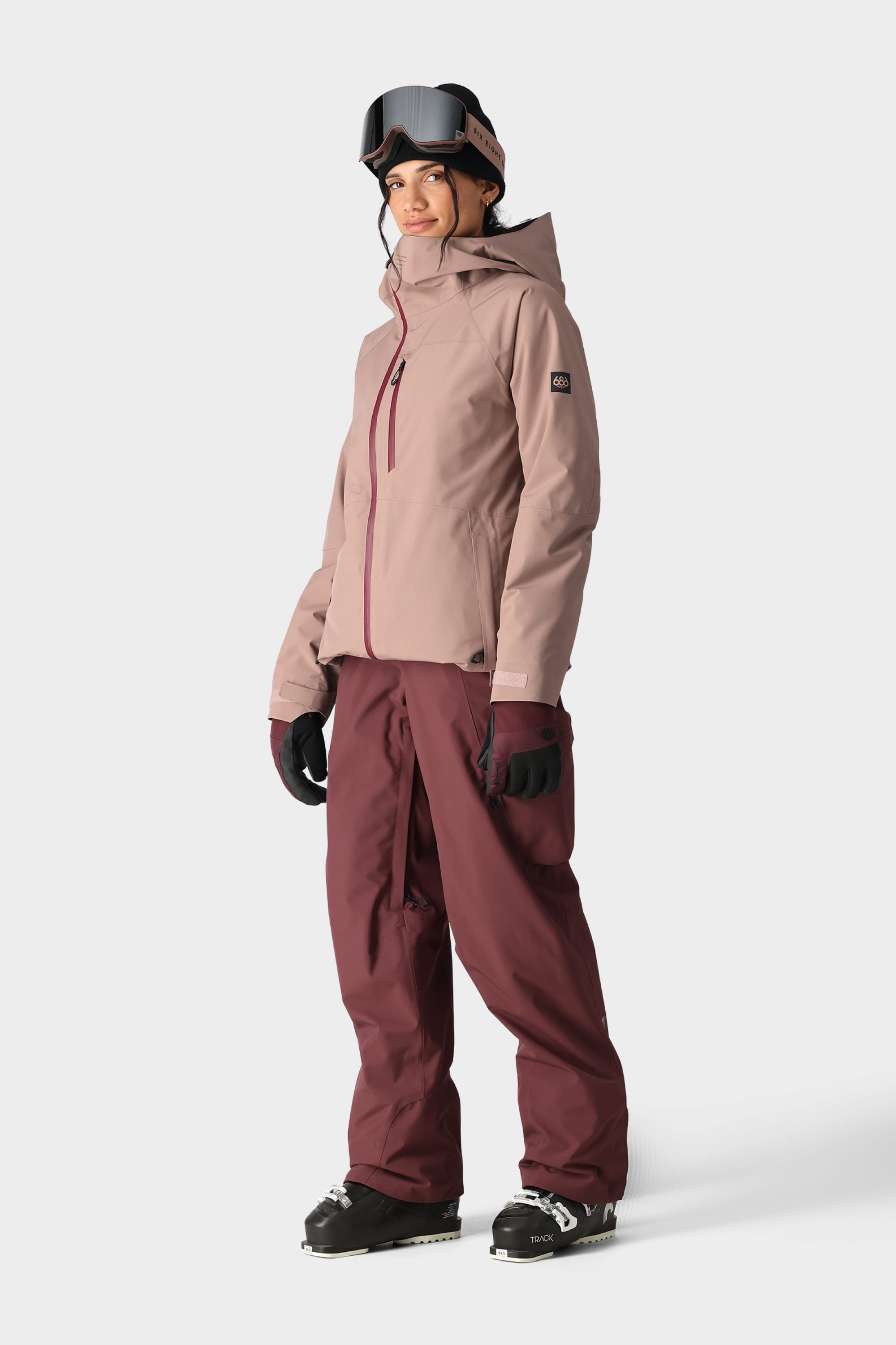 A person wears the 686 Womens Hydra Insulated Jacket (mauve, best-selling), burgundy snow pants, black gloves, ski goggles, and black ski boots while standing against a plain white background.