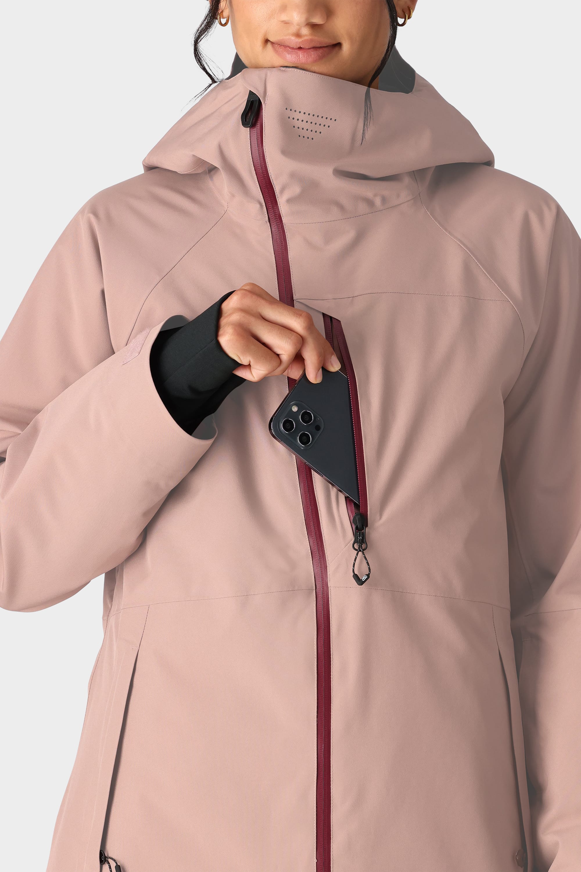 A person wearing the 686 Womens Hydra Insulated Jacket in light mauve places a black smartphone into its zippered chest pocket, with only the lower half of their face visible.