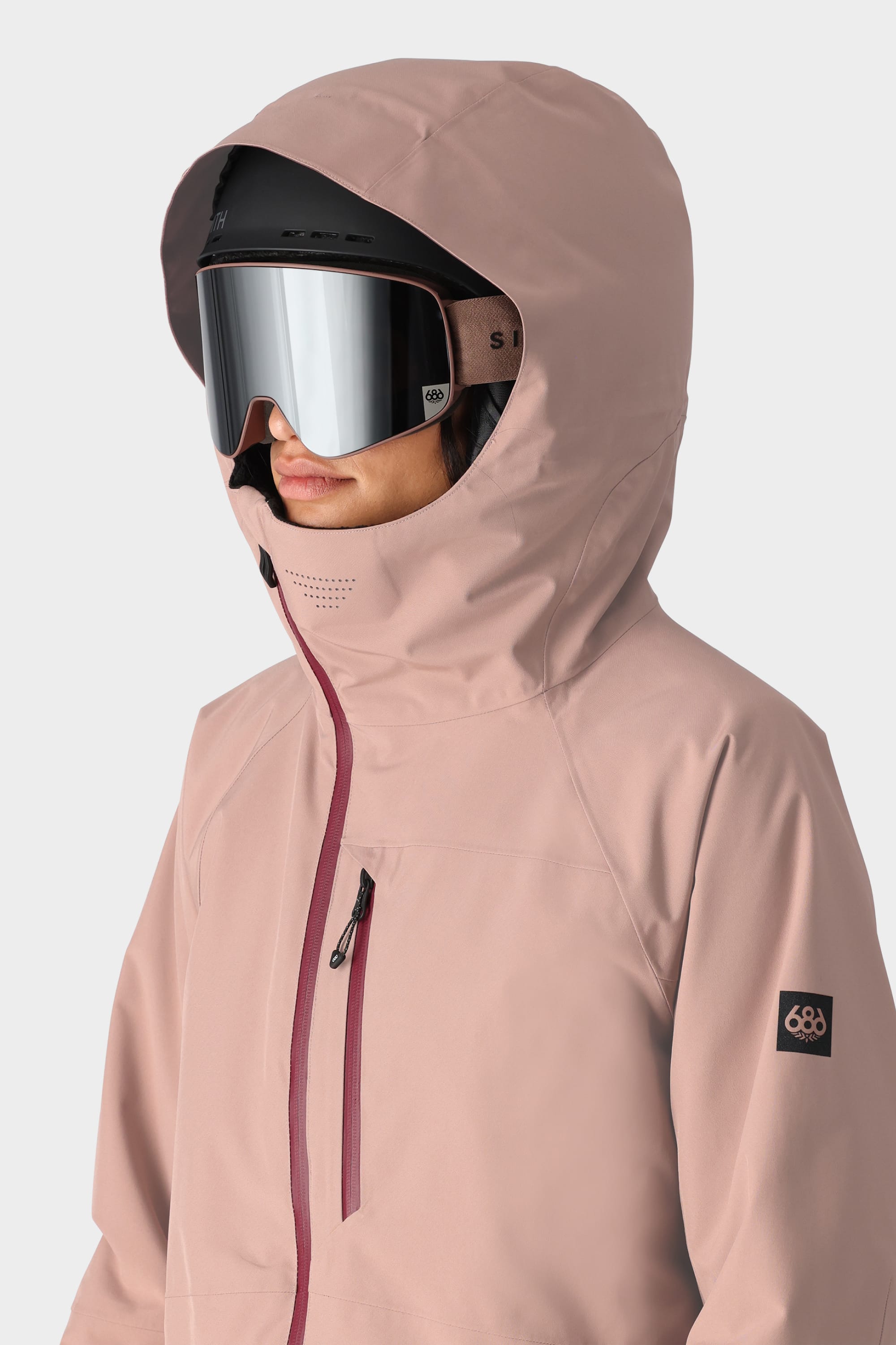 A woman wears the 686 Womens Hydra Insulated Jacket in pale pink with a maroon zipper, plus ski goggles and a helmet. The 686 logo is visible on the sleeve. Background is plain white.