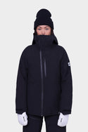 A person wearing the 686 Womens Hydra Insulated Jacket in black with light insulation, a high collar, a black beanie, and white gloves stands facing forward against a plain white background.