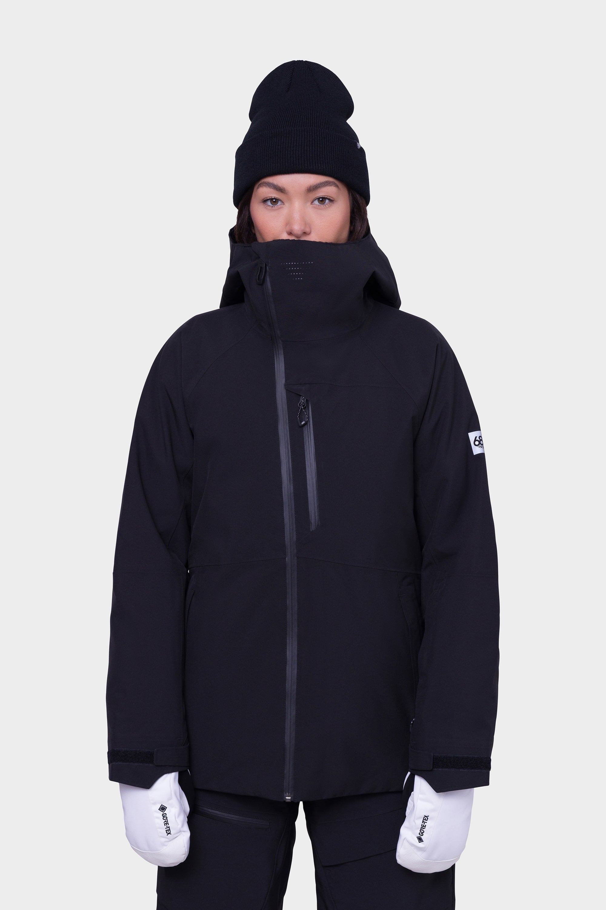 A person wearing the 686 Womens Hydra Insulated Jacket in black with light insulation, a high collar, a black beanie, and white gloves stands facing forward against a plain white background.