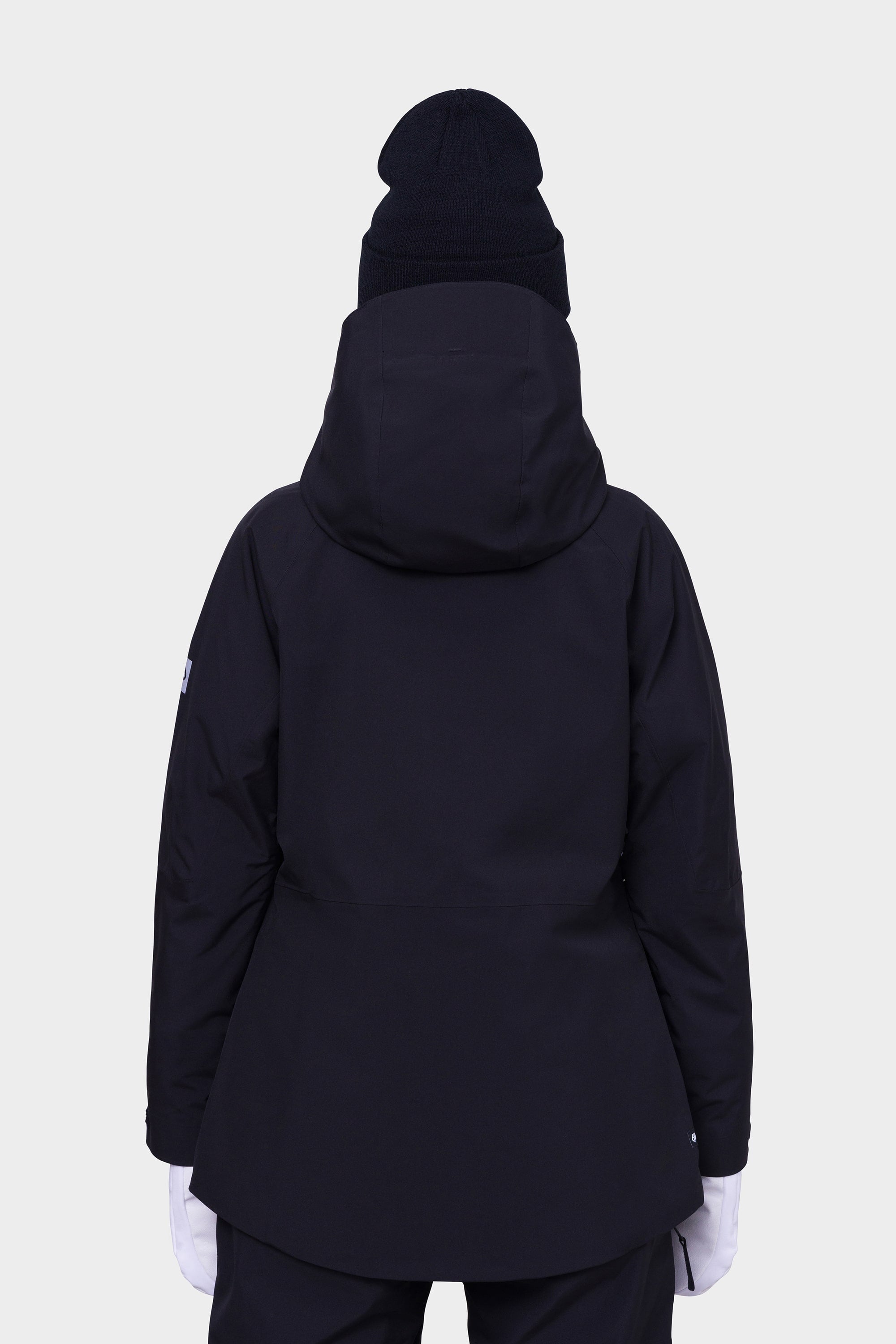 A person wearing a 686 Womens Hydra Insulated Jacket in black, paired with a black beanie and white gloves, shown from behind against a plain light background.
