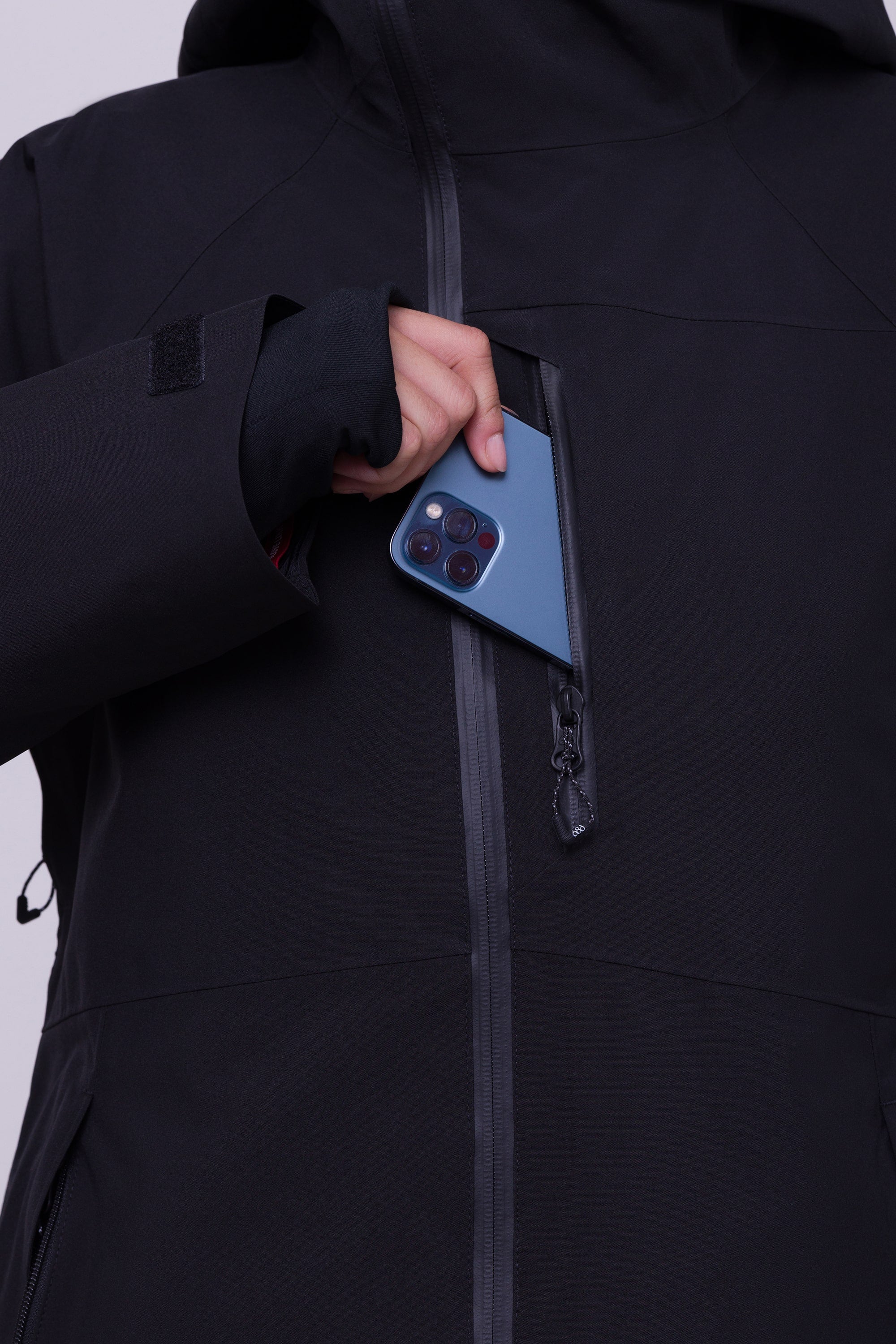 A woman wearing the 686 Womens Hydra Insulated Jacket slips a blue smartphone into its partially unzipped chest pocket; only her hand, phone, and upper body are visible.