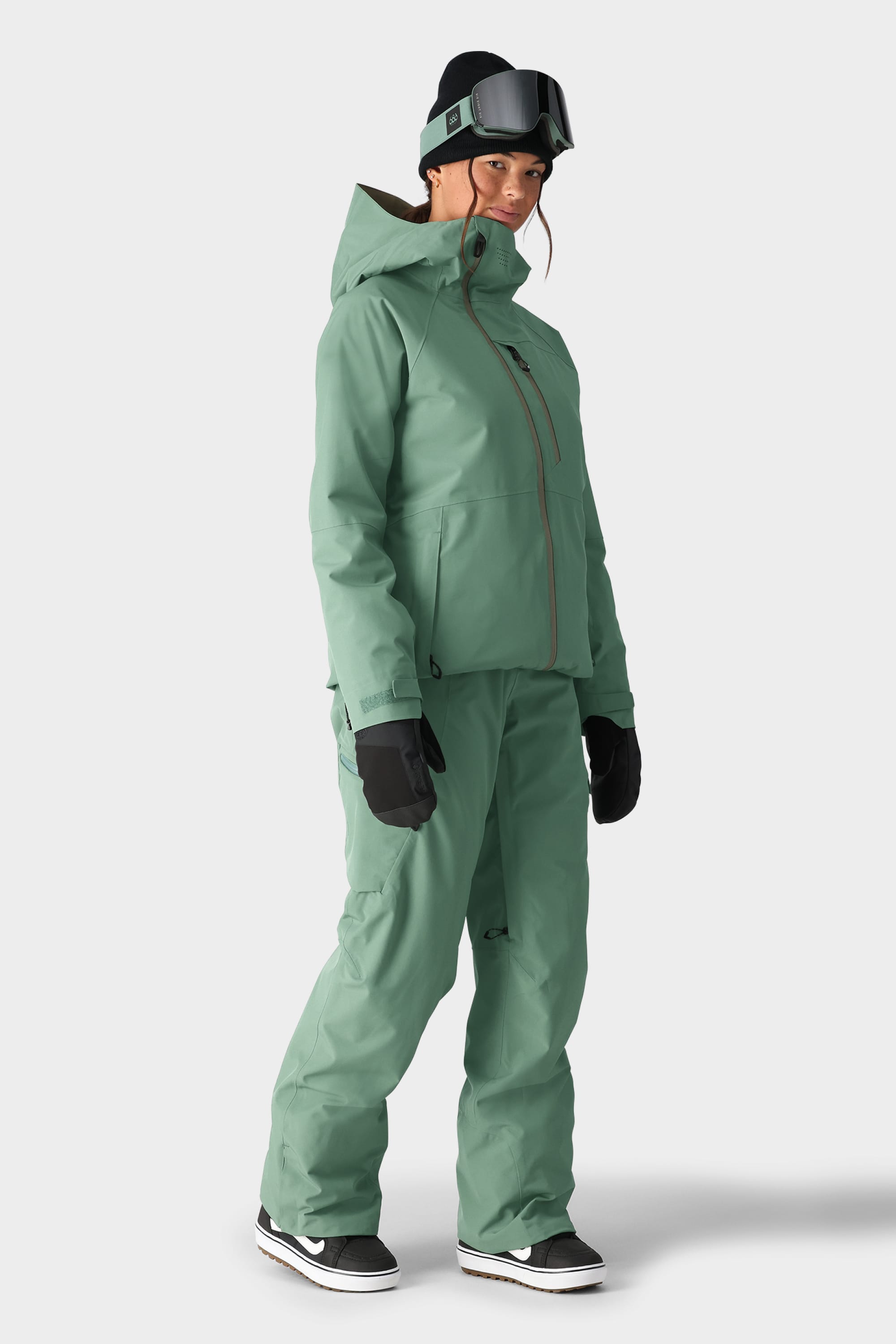 A person wears the 686 Womens Hydra Insulated Jacket in sage green with matching pants, black gloves, a black beanie, ski goggles on their forehead, and black and white sneakers against a plain light background.