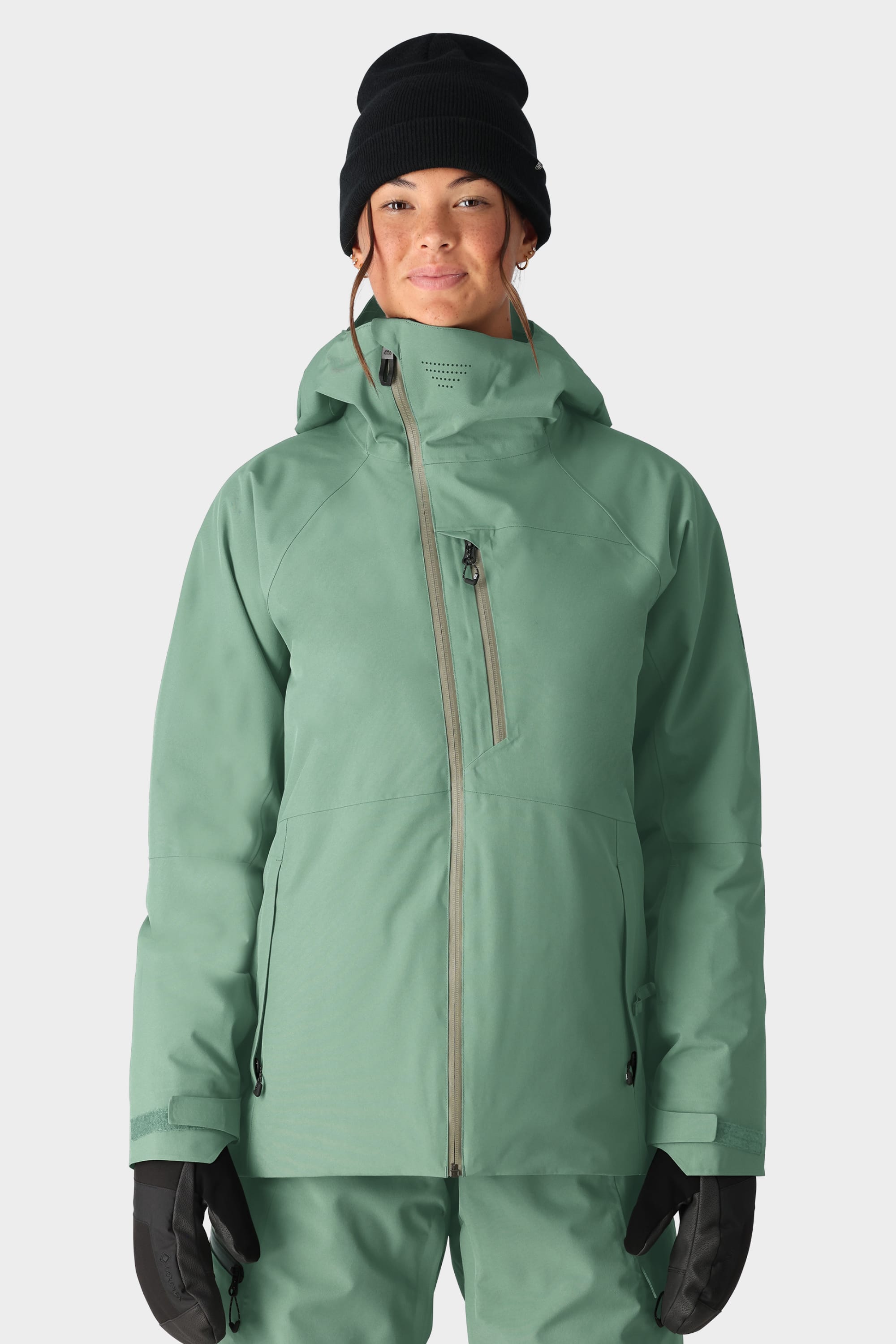 A person wearing the 686 Womens Hydra Insulated Jacket in mint green, with matching pants, black gloves, and a black beanie stands facing forward against a plain light background.