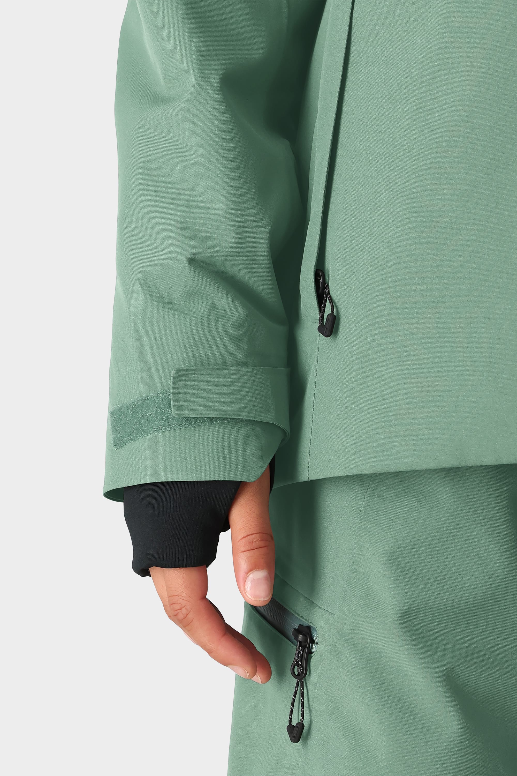 A person wears the 686 Womens Hydra Insulated Jacket in light green with matching pants, unzipping a pant pocket. The jacket sleeve has a velcro strap and partially covers a black glove.
