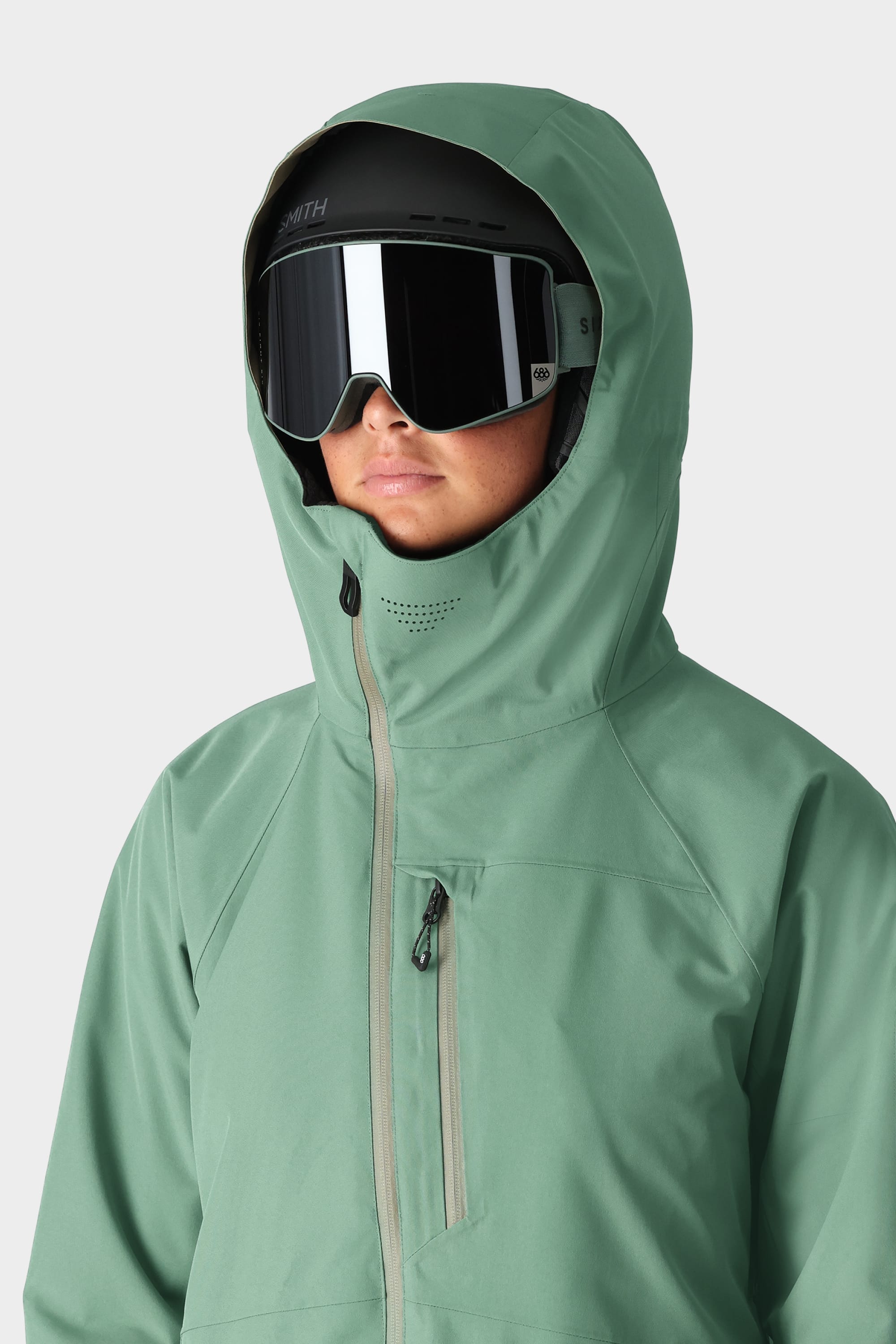A person wears a 686 Womens Hydra Insulated Jacket in green, zipped up to cover most of their face, along with black ski goggles and a black helmet. The background is plain white.