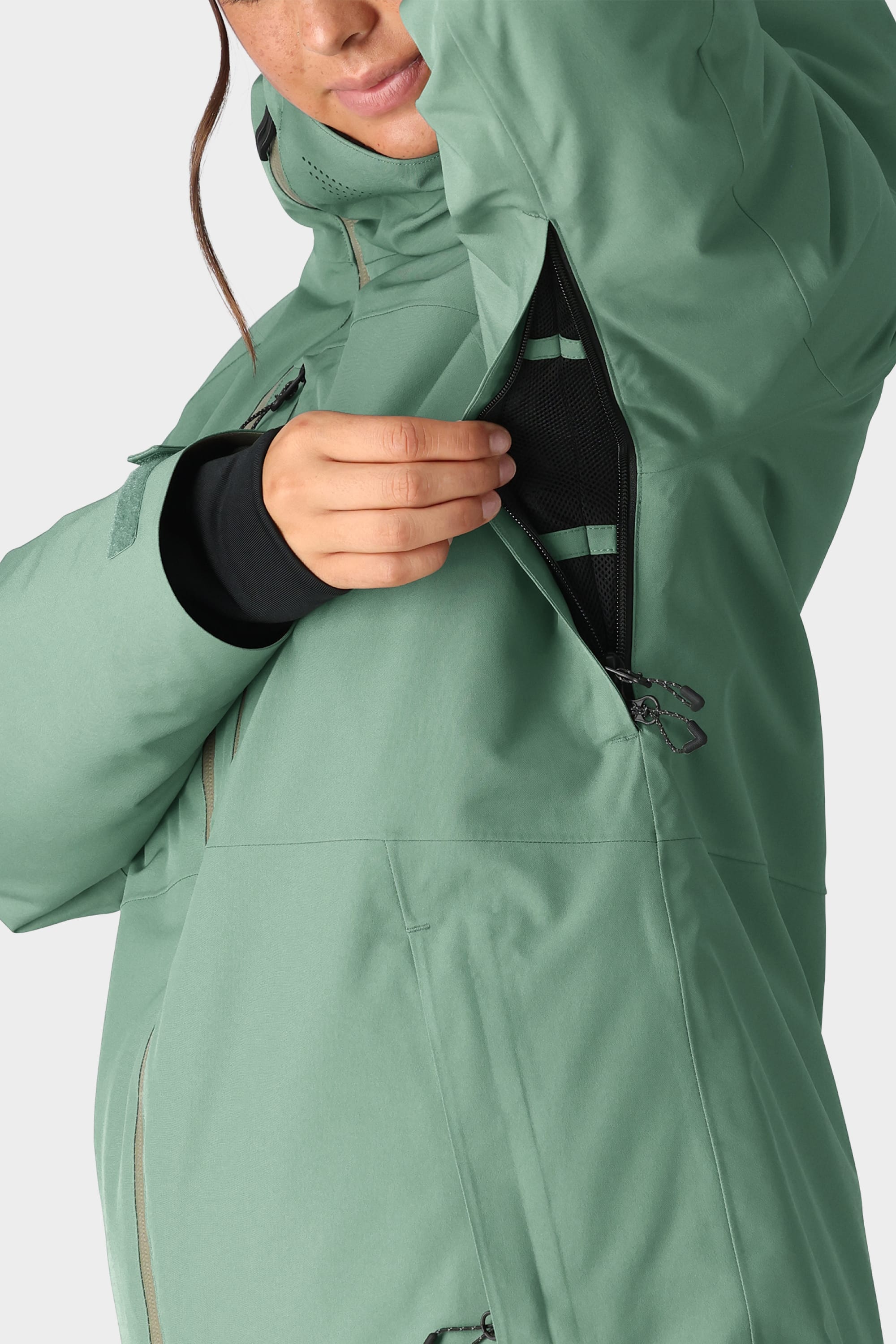 A woman in a light green 686 Womens Hydra Insulated Jacket, a best-seller with light insulation, unzips a black mesh-lined underarm vent. Only the lower part of her face and upper body are visible.