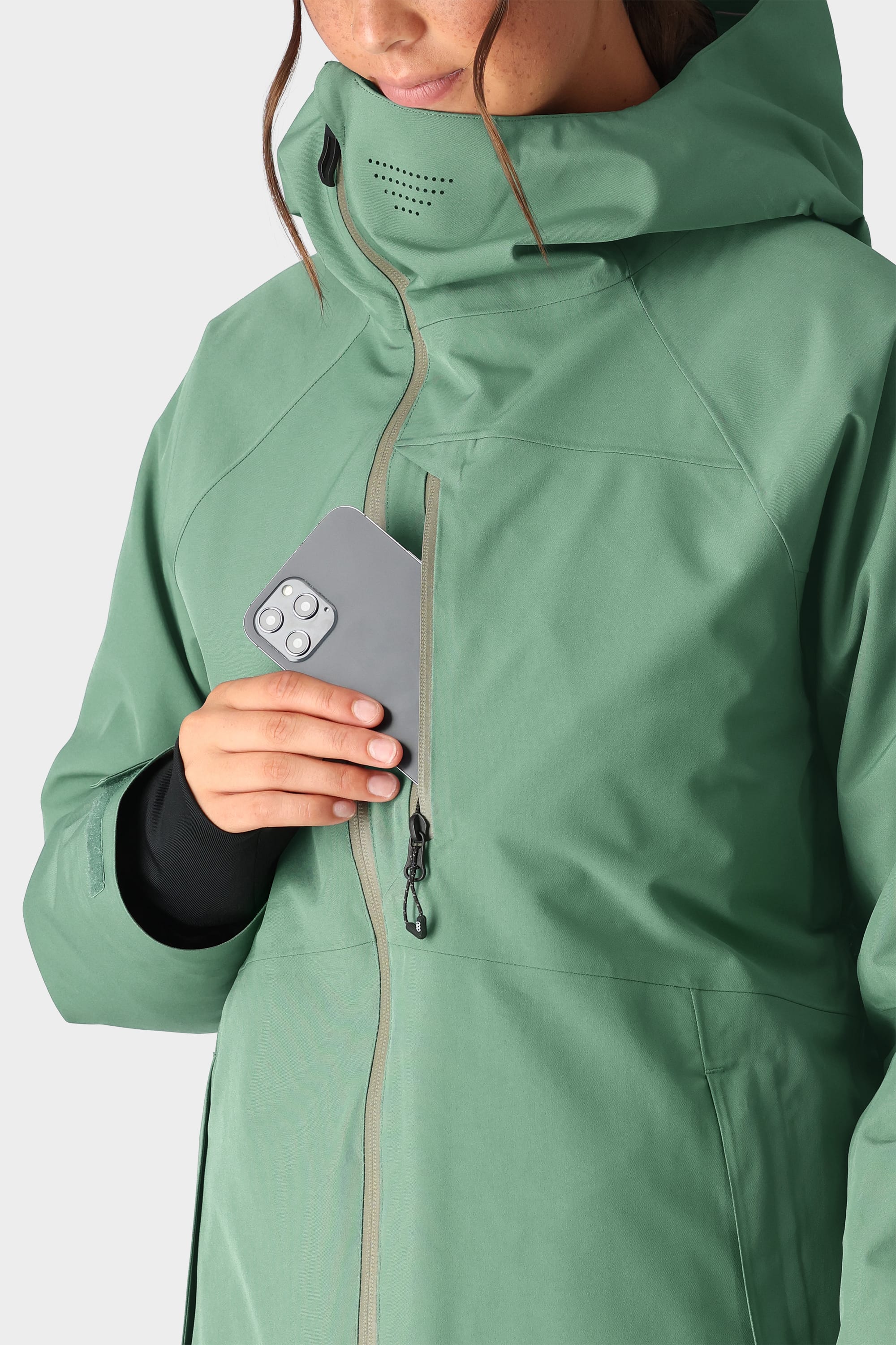 A person wearing the 686 Womens Hydra Insulated Jacket, a green waterproof piece with light insulation and hood, slips a silver smartphone into its zippered chest pocket. The jacket features laser-cut ventilation holes near the collar.