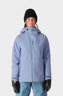 A person wearing a black beanie, the 686 Womens Hydra Insulated Jacket in light purple, blue snow pants, and black gloves stands facing forward against a plain, light background.