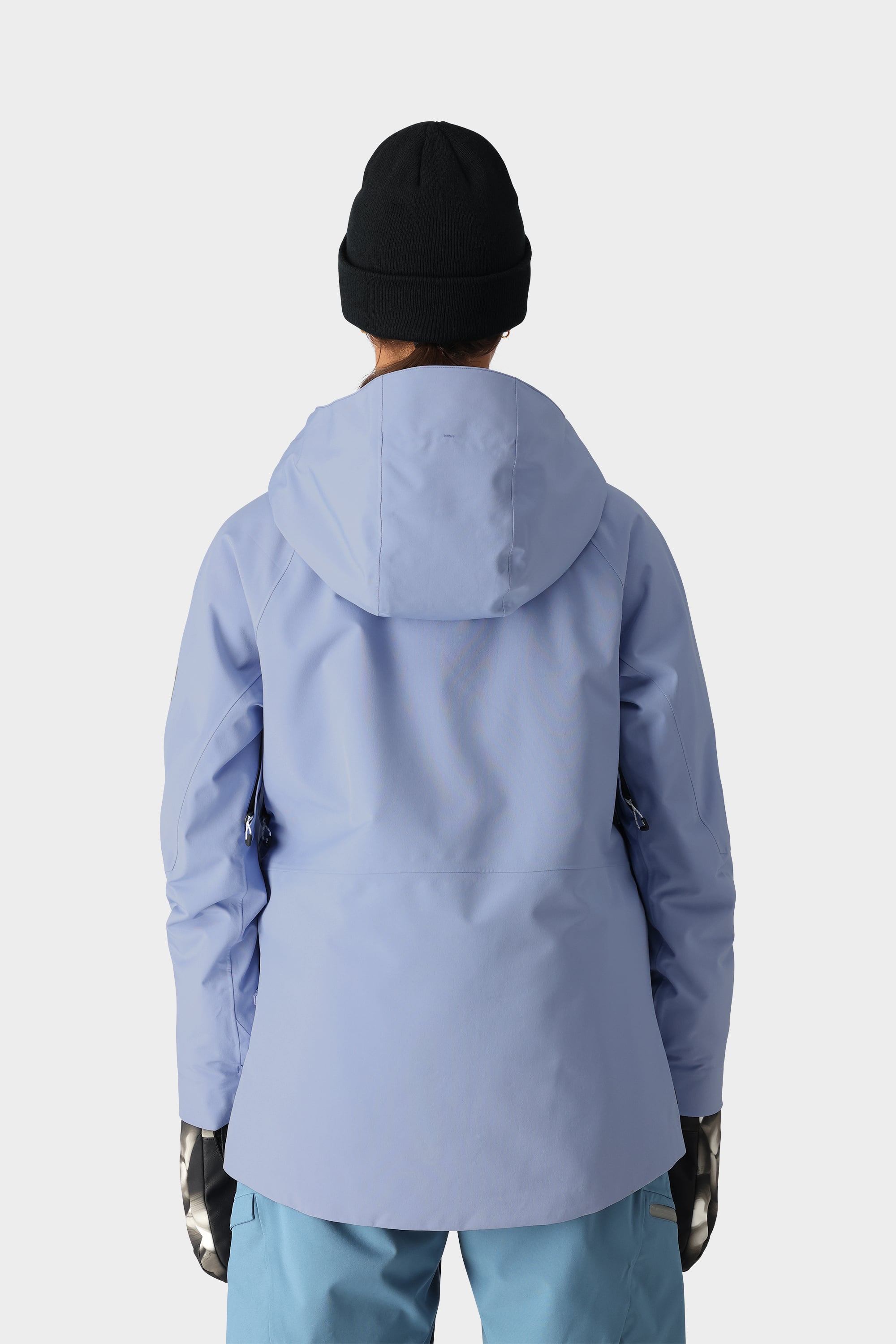 A person is seen from behind wearing the best-selling 686 Womens Hydra Insulated Jacket in light purple, paired with blue pants, black beanie, and patterned gloves, against a plain white background.