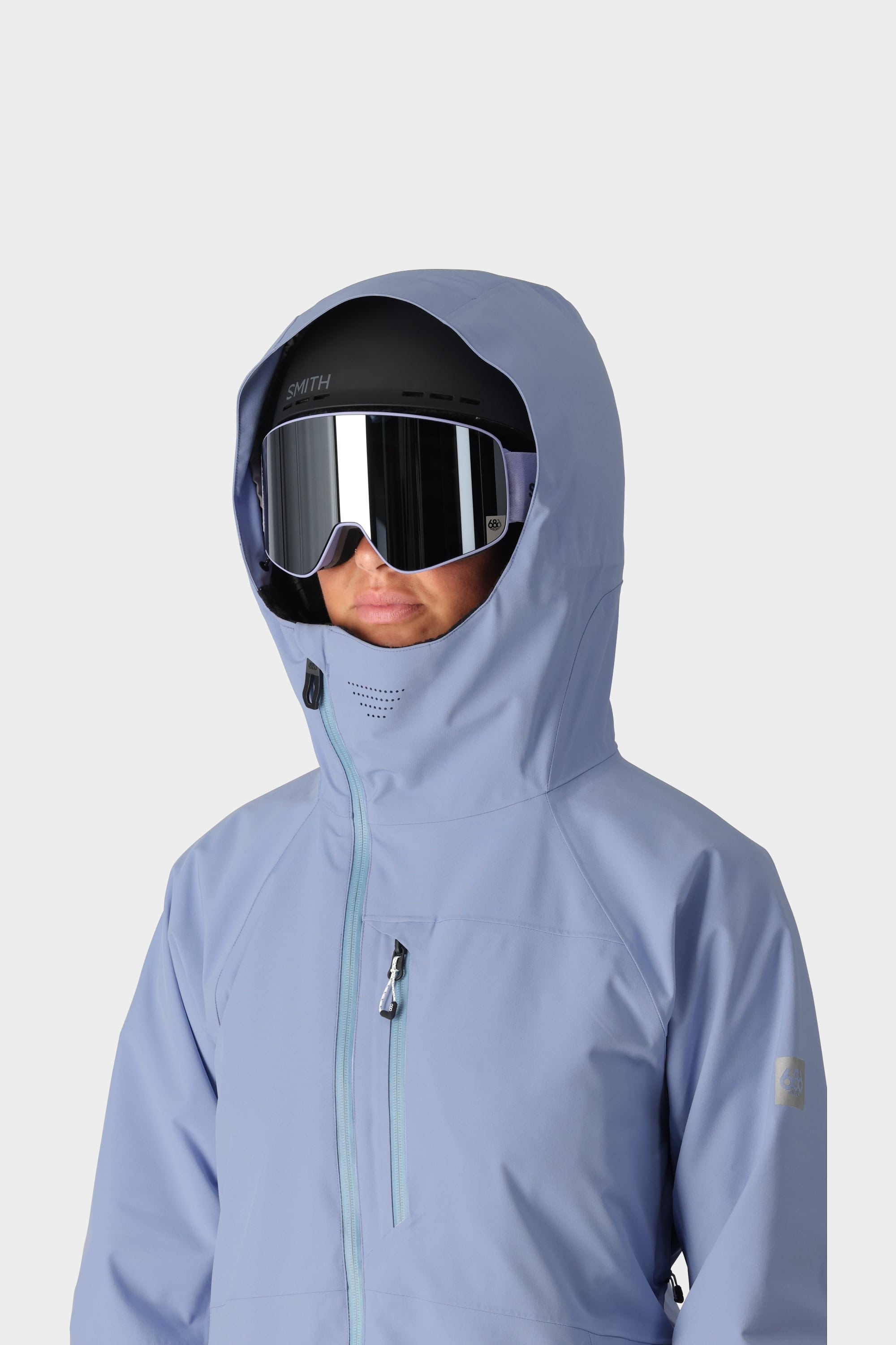 A person wears the 686 Womens Hydra Insulated Jacket in light blue, paired with a black ski helmet and reflective ski goggles, hood up, against a plain light background.