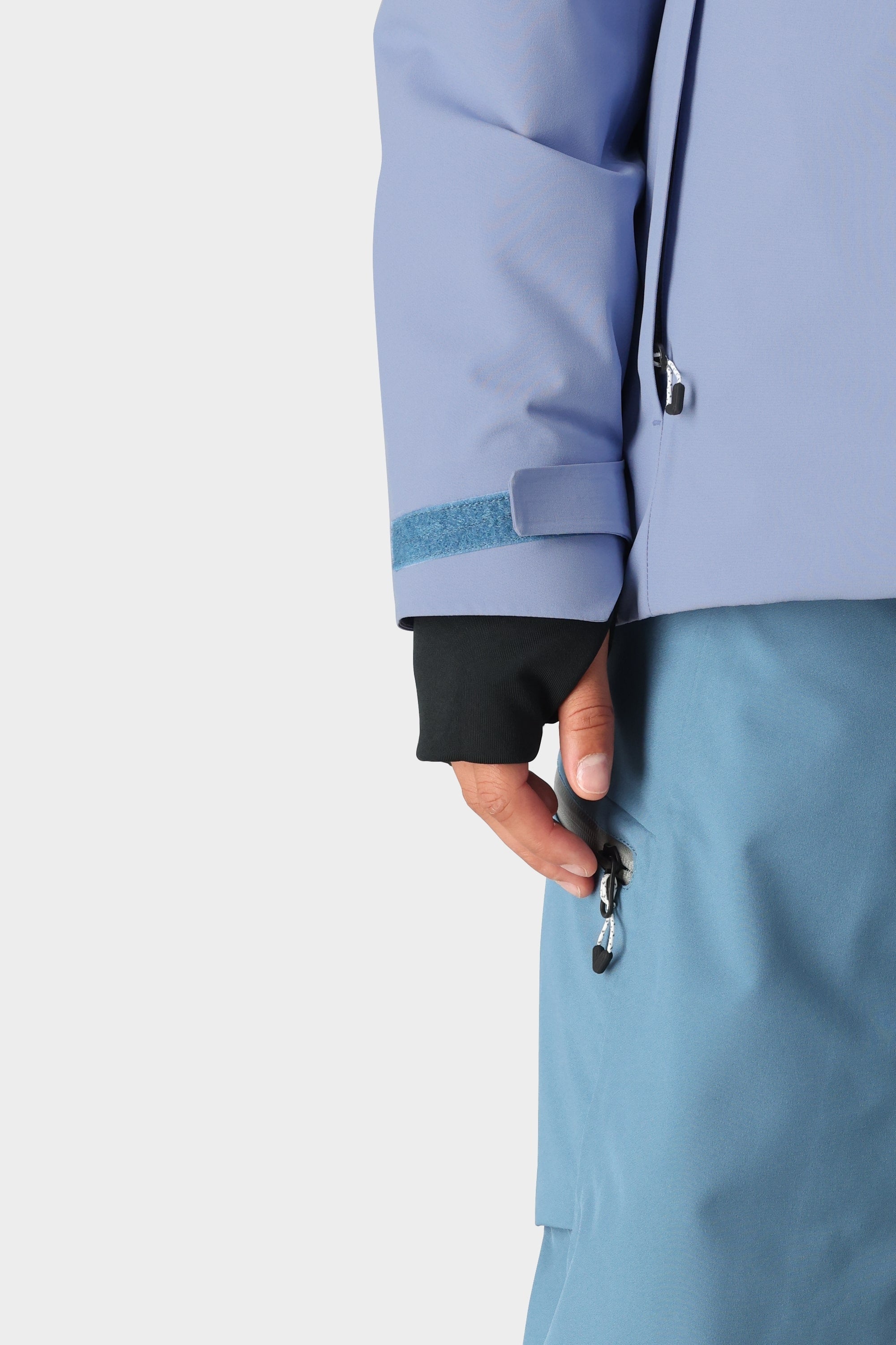A close-up of a person wearing the 686 Womens Hydra Insulated Jacket in light blue, featuring light insulation, a velcro strap on the sleeve, and a zippered pocket, paired with blue pants. One hand is partially visible near the pocket.