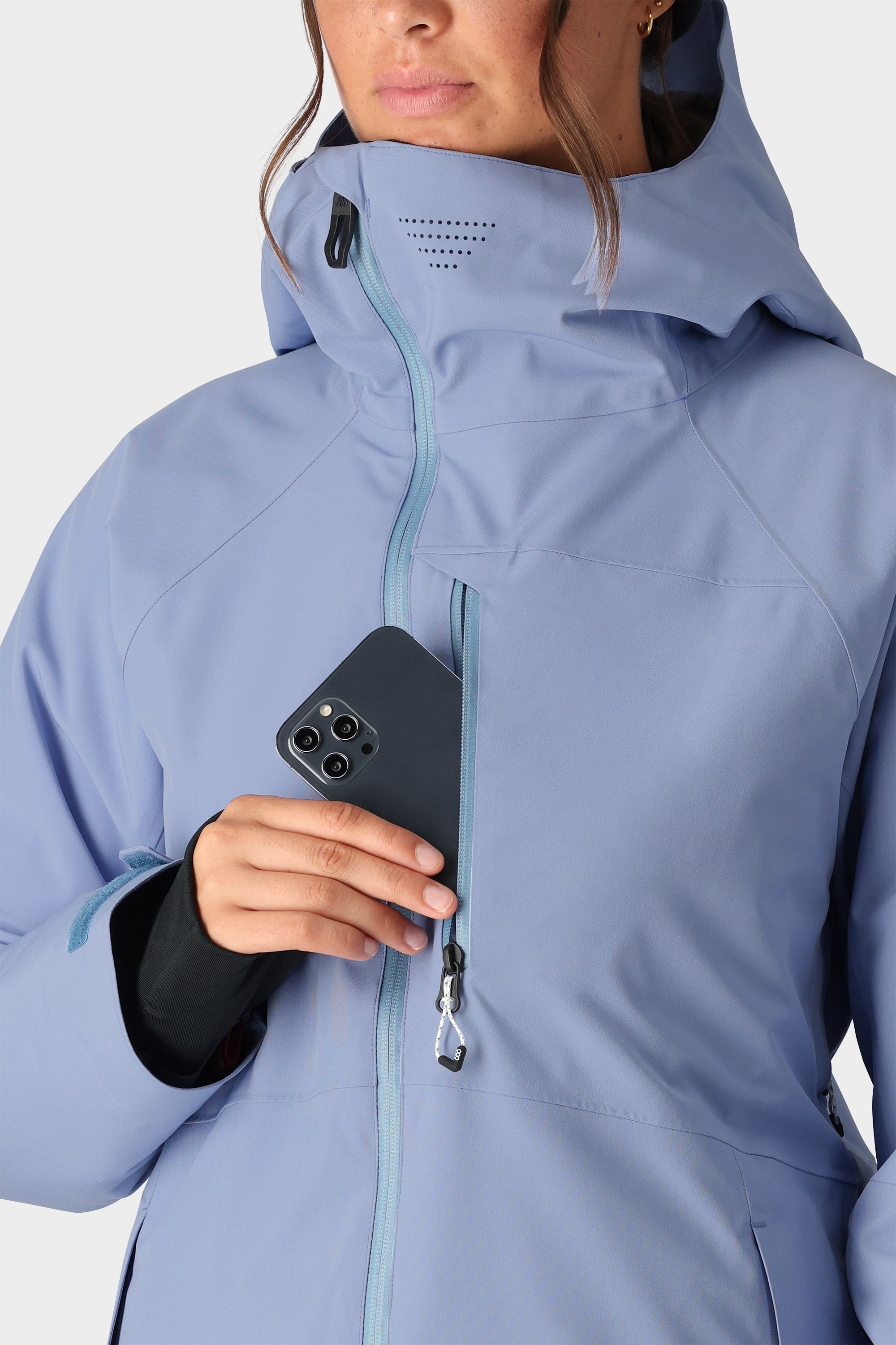 Wearing the 686 Womens Hydra Insulated Jacket in light blue, a woman places a black smartphone into its zippered chest pocket, with only her lower face and upper body visible.