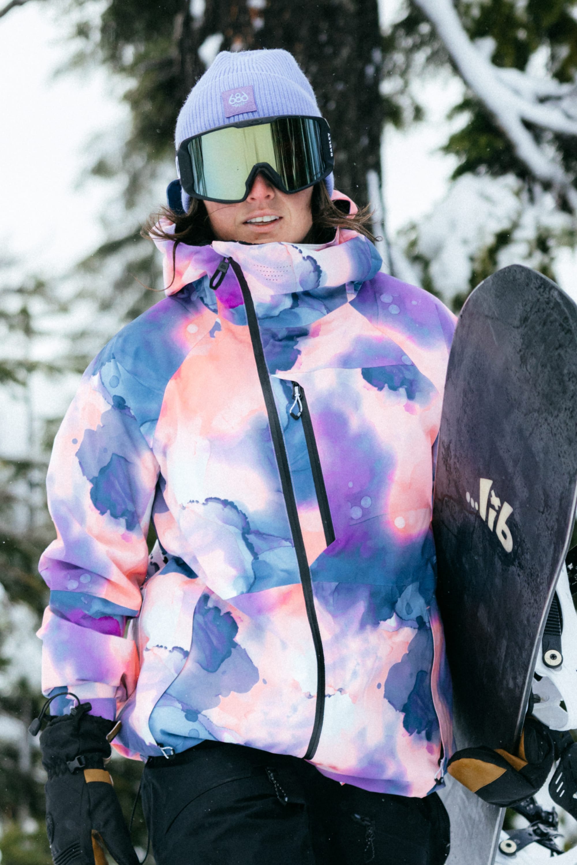 A snowboarder in a best-selling 686 Womens Hydra Insulated Jacket with pastel tie-dye pattern, light insulation, purple beanie, and reflective goggles stands holding a snowboard in a snowy forest.
