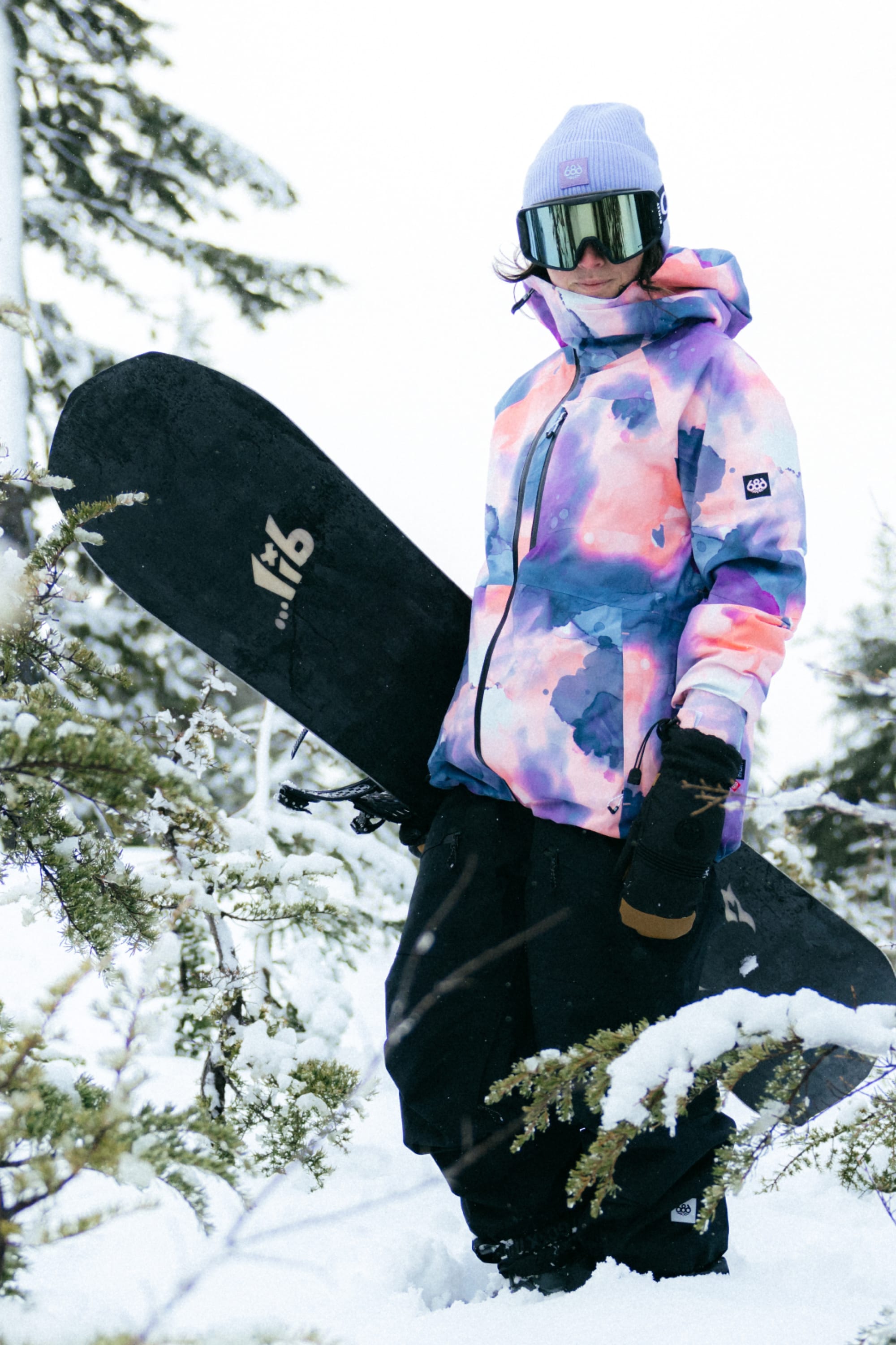 Wearing the 686 Womens Hydra Insulated Jacket, black pants, gloves, a light purple beanie, and ski goggles, a person stands in the snow with a snowboard amid snow-covered trees.