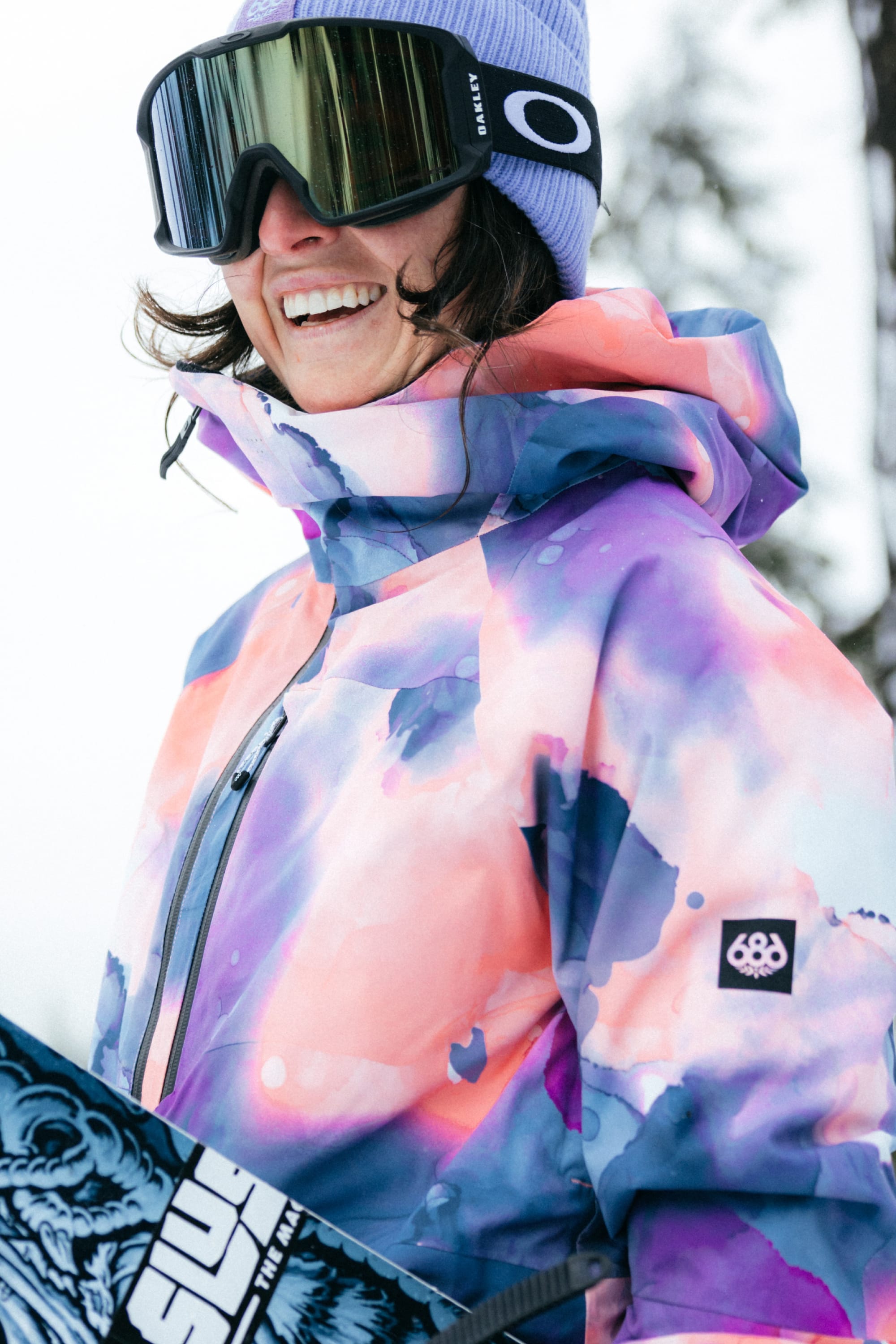 A woman in the 686 Womens Hydra Insulated Jacket, a best-seller from 686, smiles holding a snowboard. She wears a purple beanie and large ski goggles, with snowy trees softly blurred in the background.