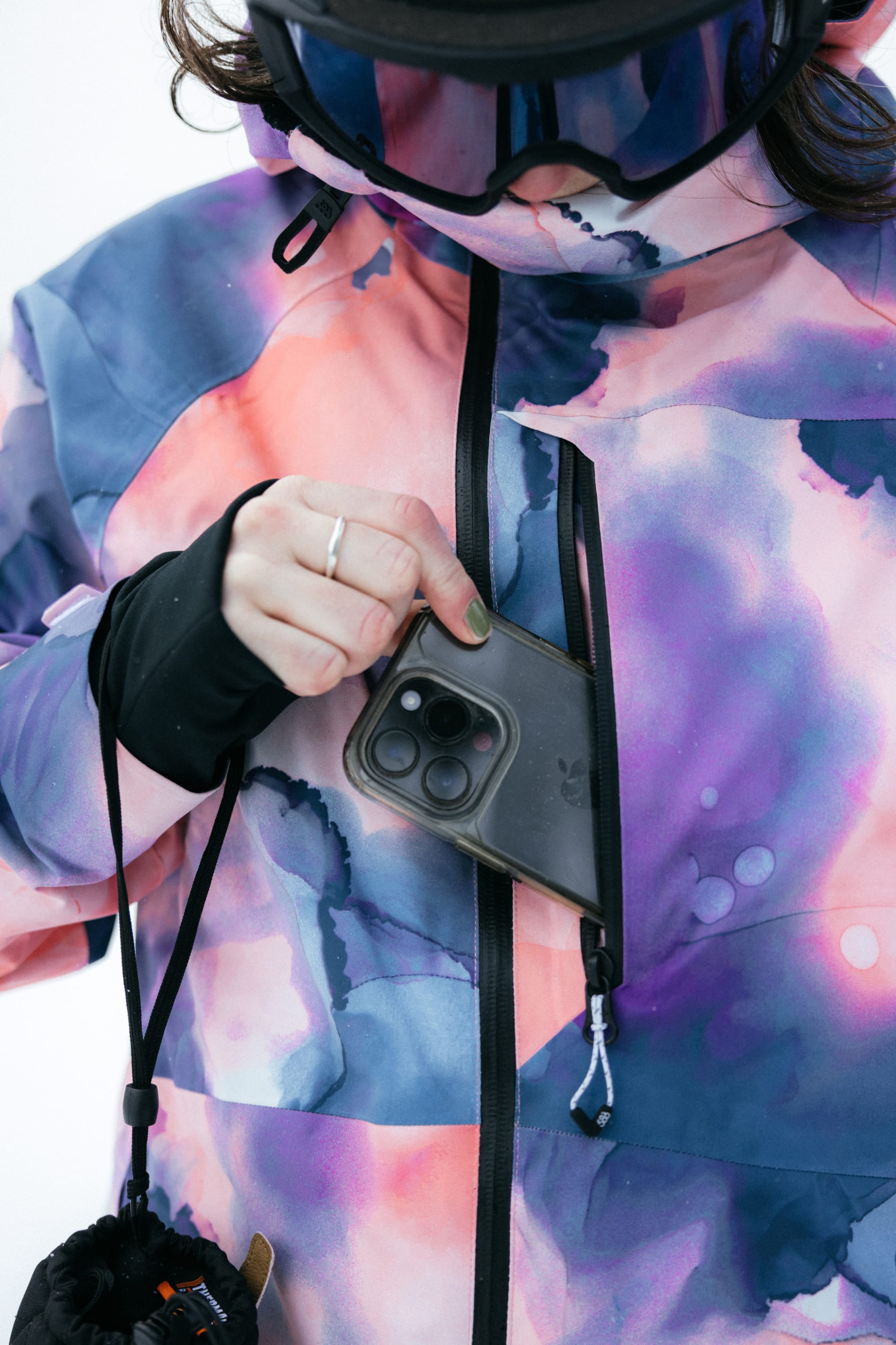 Wearing the 686 Womens Hydra Insulated Jacket in a colorful watercolor pattern, a person puts a smartphone into its zippered chest pocket. They’re also outfitted with black gloves and ski goggles.