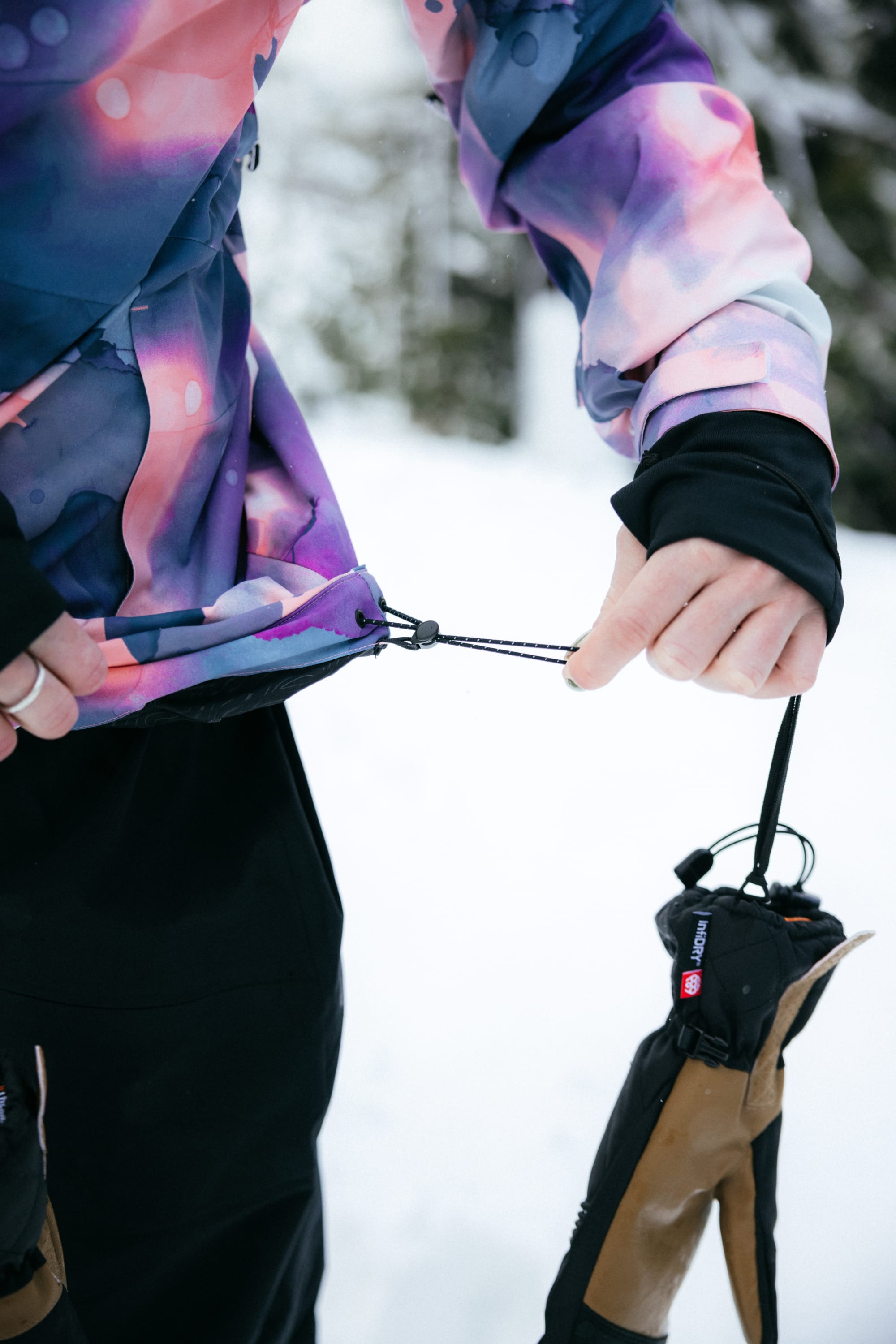 A person wearing the 686 Womens Hydra Insulated Jacket tightens the hem drawstring while holding gloves in a snowy outdoor setting.