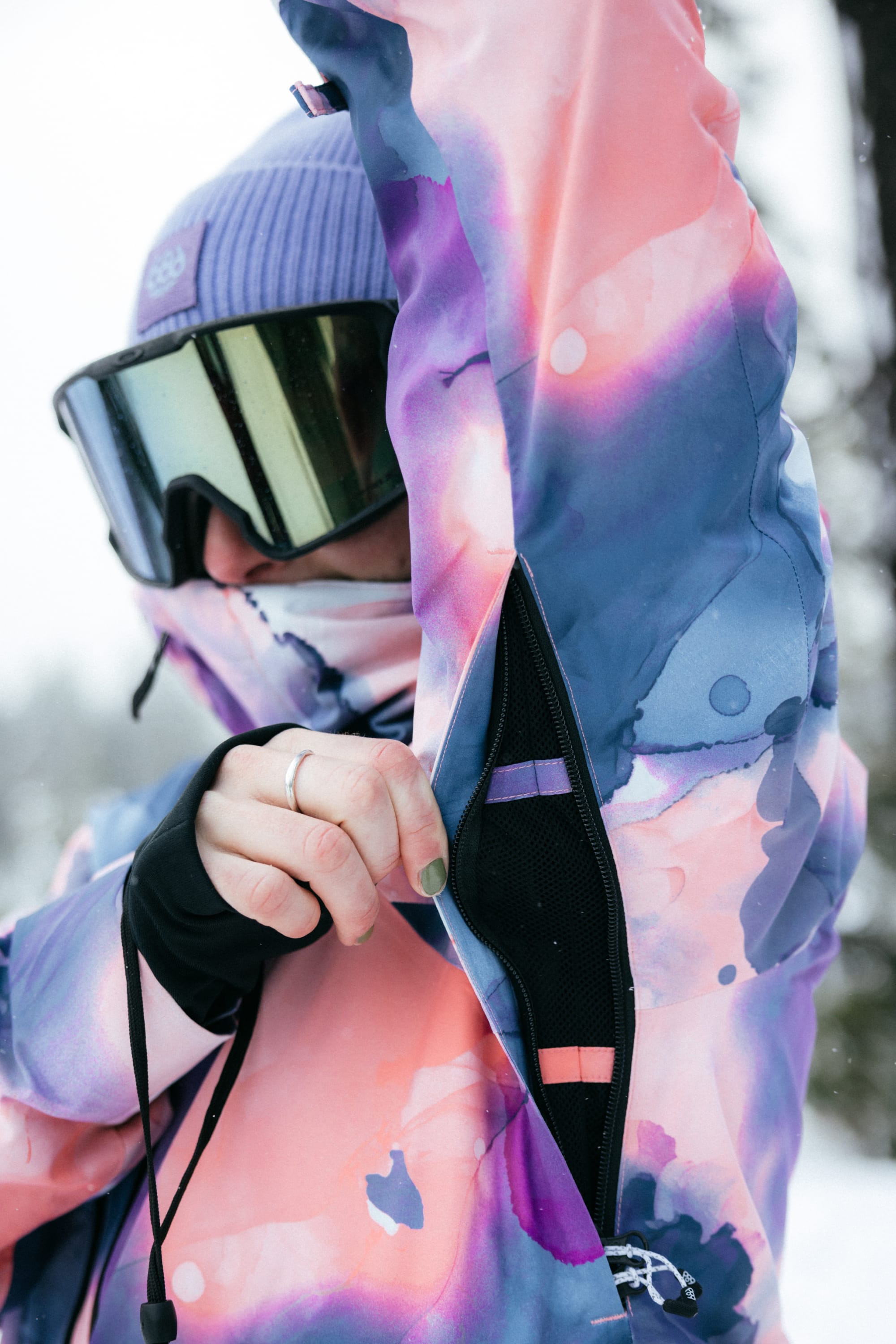 Wearing the 686 Womens Hydra Insulated Jacket—a colorful, waterproof coat with light insulation—a person in mirrored goggles opens a zippered underarm vent, with snow and trees blurred in the background.