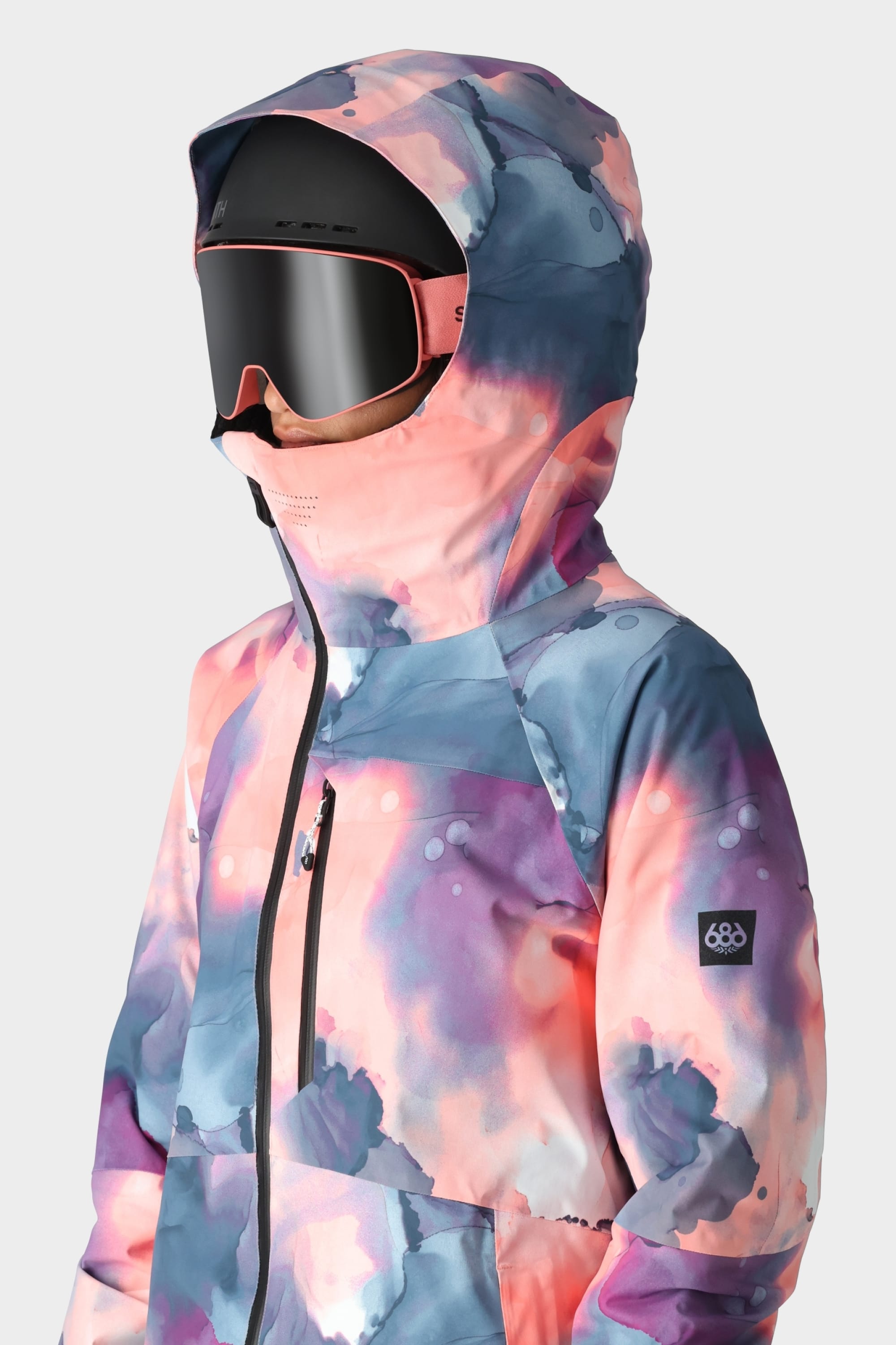 A person wearing the 686 Womens Hydra Insulated Jacket in a pink, purple, and blue watercolor pattern with a hood, paired with large black ski goggles and a face covering. The background is plain light gray. Brand: 686.