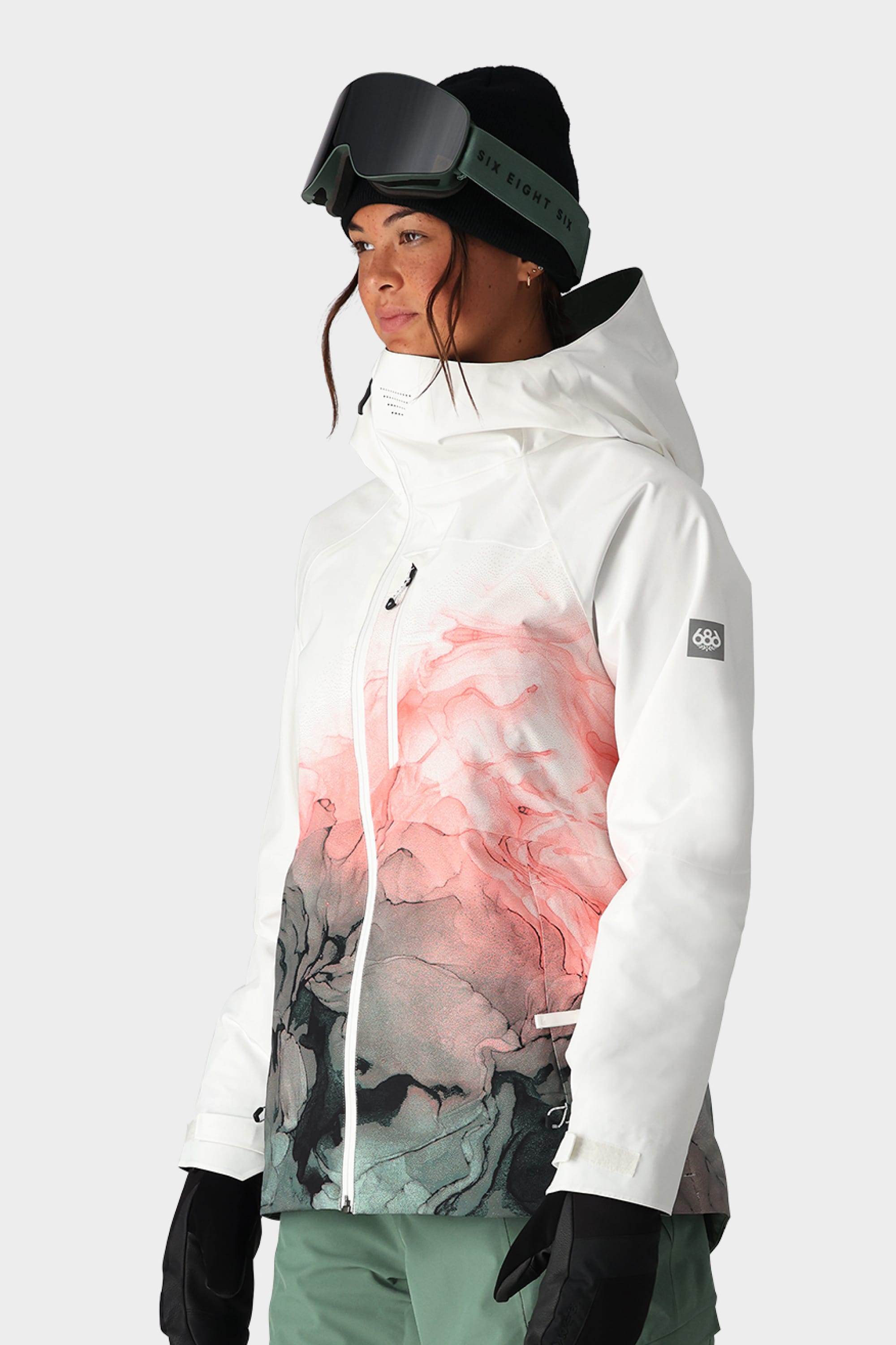 A woman wears the 686 Womens Hydra Insulated Jacket by 686—white with pink and gray abstract patterns—paired with green ski pants, black gloves, a black beanie, and ski goggles on her head against a light gray background.