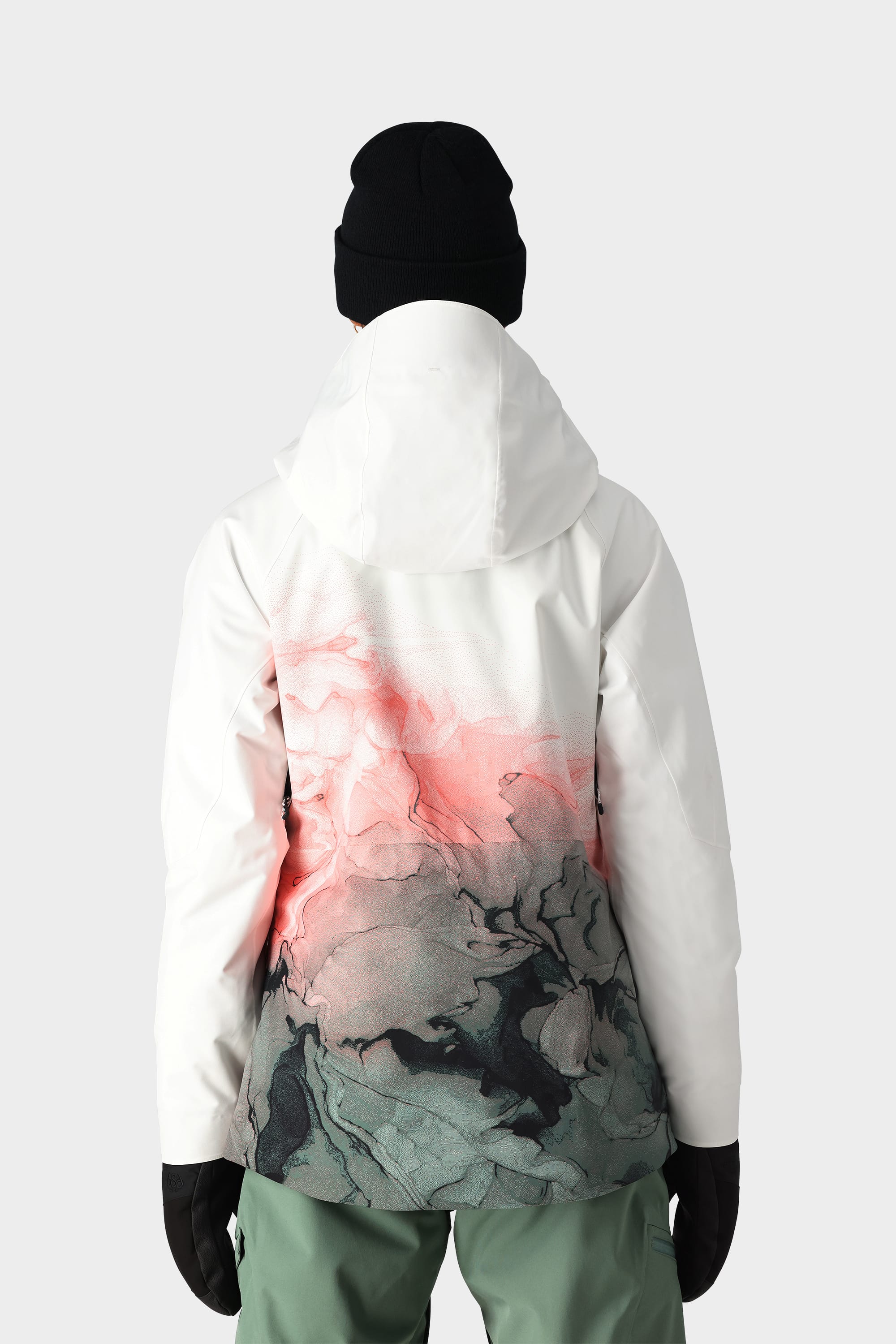 Wearing the 686 Womens Hydra Insulated Jacket by 686 with a pink and dark gray abstract back pattern, light insulation for comfort, green pants, black gloves, and a black beanie against a plain light background.