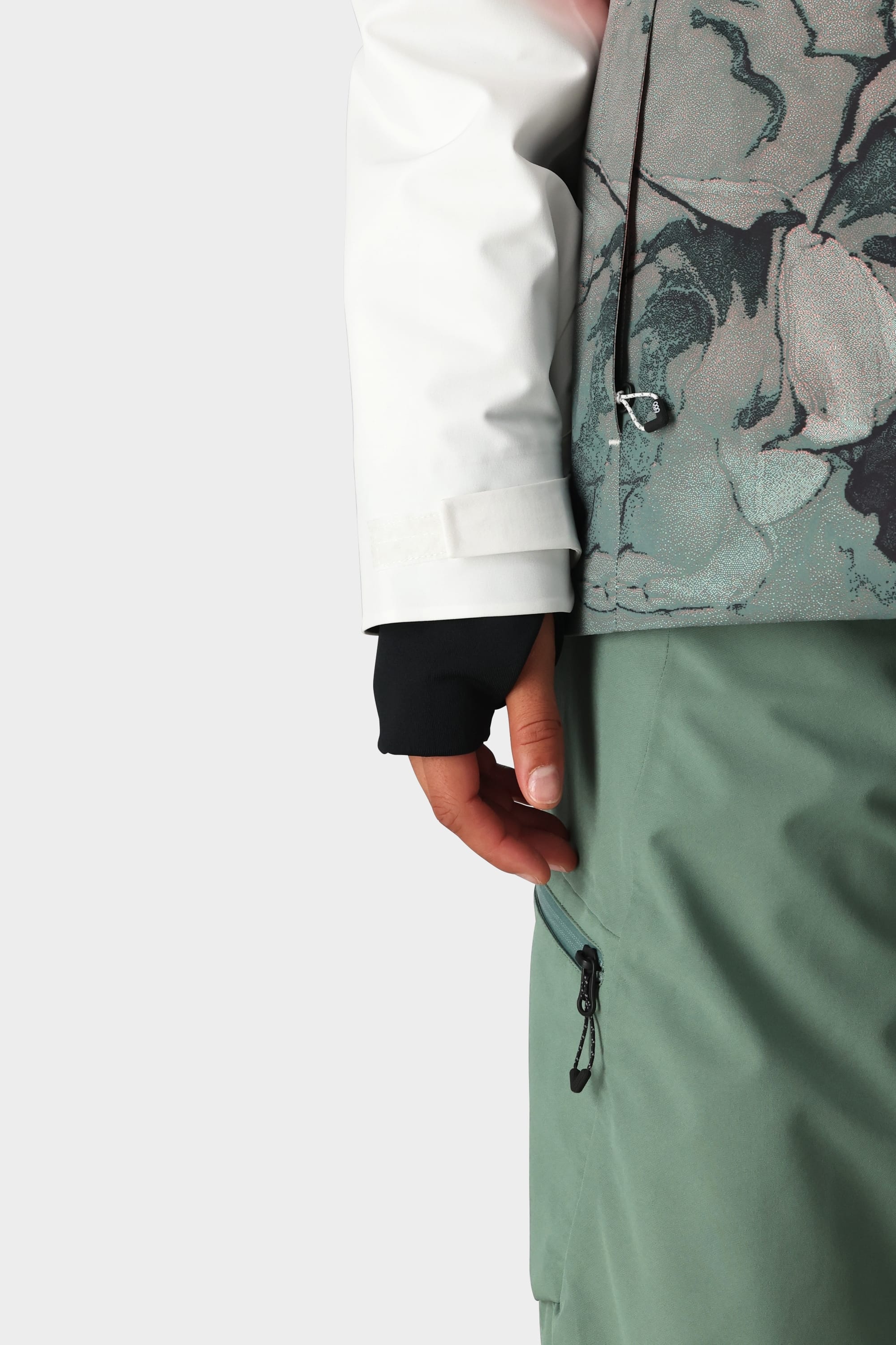 A person wearing the 686 Womens Hydra Insulated Jacket with floral waterproof sleeves, black gloves, and green pants with a zippered pocket holds the zipper against a plain background.