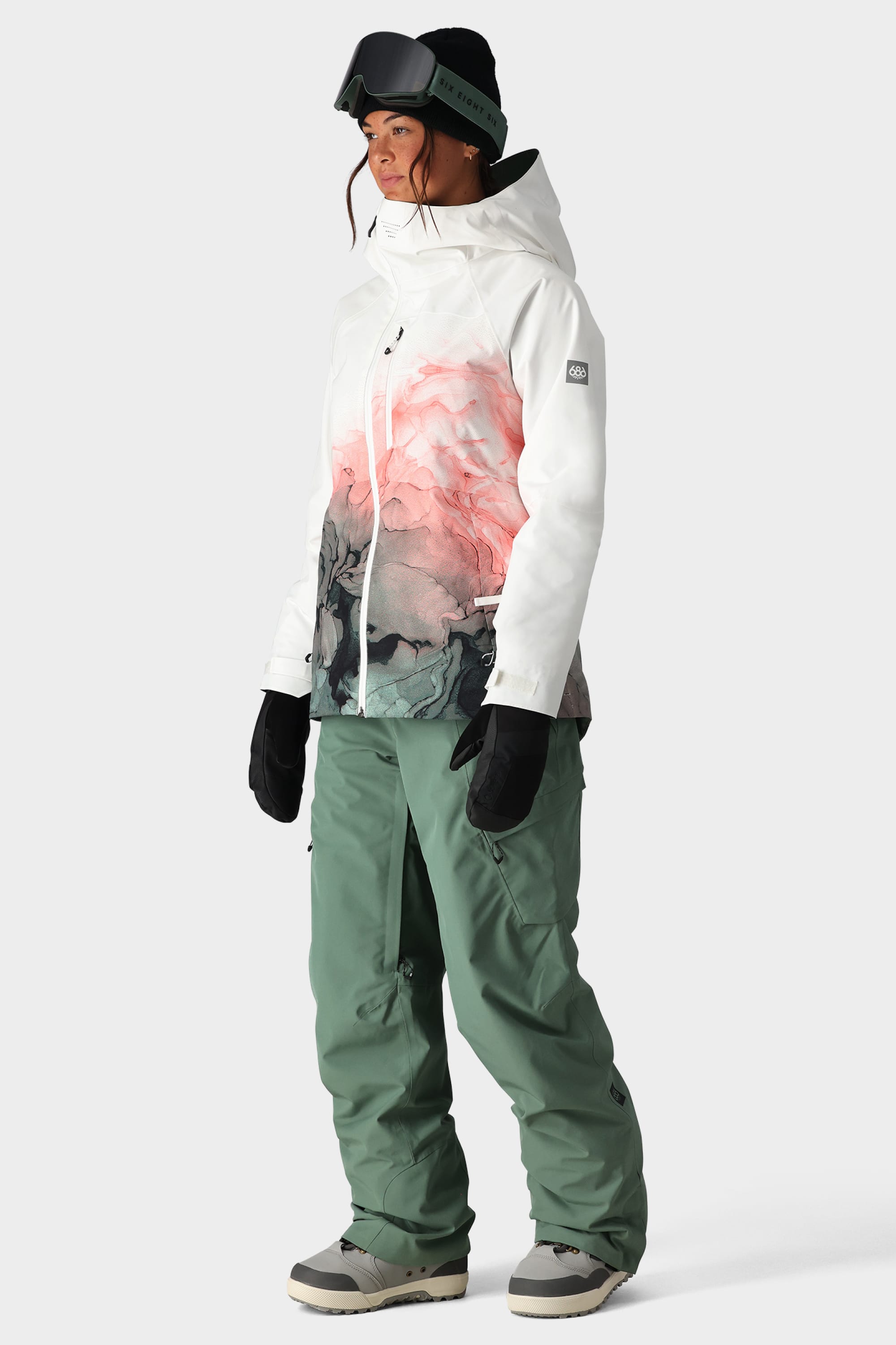 A woman wearing the 686 Womens Hydra Insulated Jacket, green snow pants, black gloves, beige boots, and black ski goggles stands slightly facing left against a plain background.