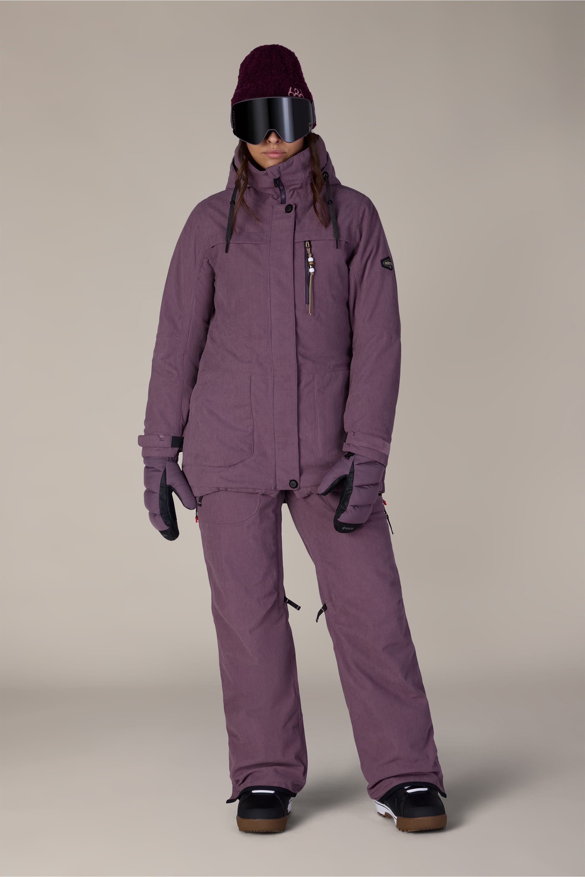 A person wearing the 686 Women’s Spirit Insulated Jacket in purple, paired with matching ski pants, black gloves, dark snow goggles, and a maroon beanie stands against a plain light background.